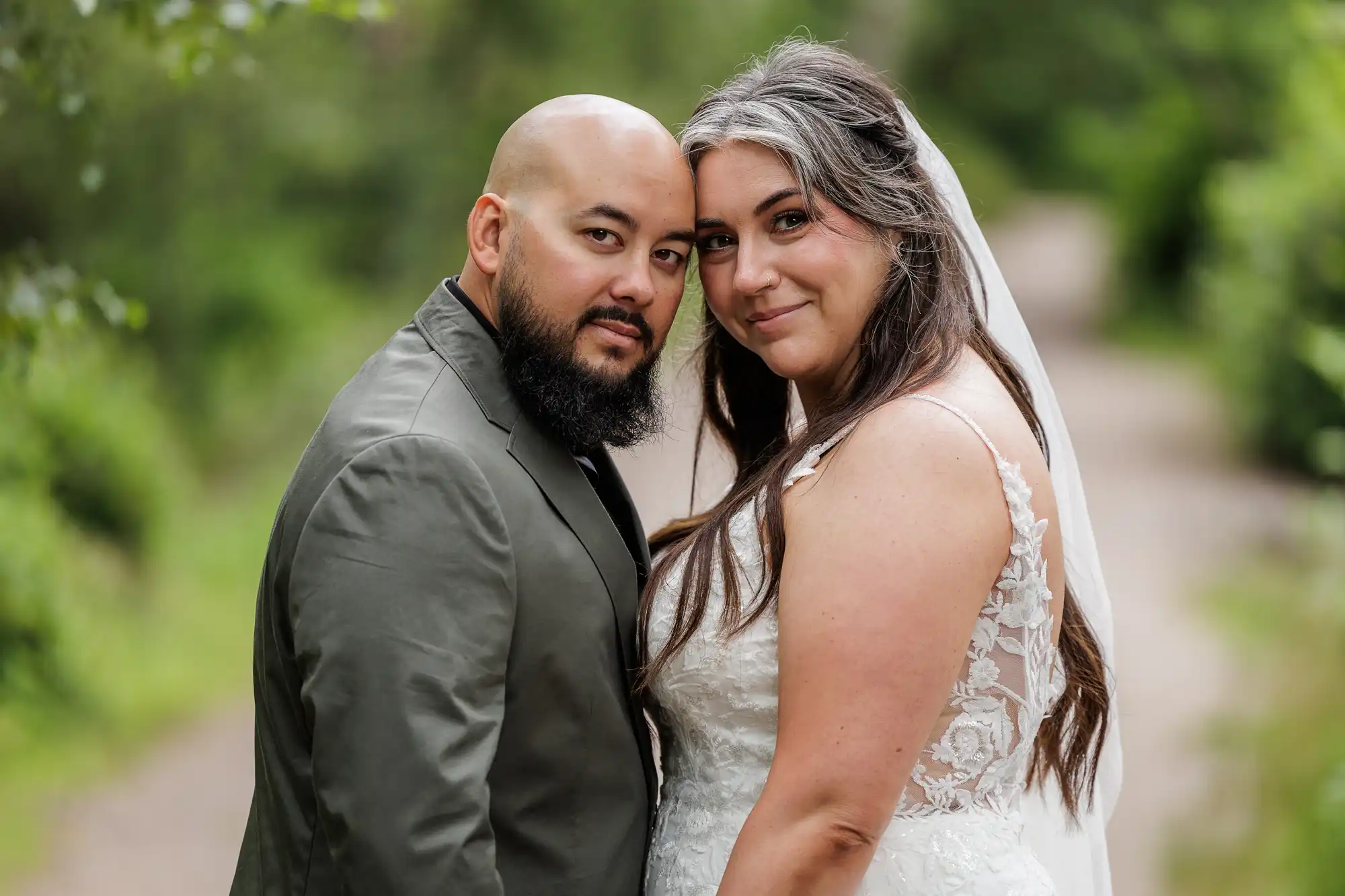 A couple pose outdoors on a leafy path. The man wears a dark suit and has a beard and shaven head. The woman wears a white lace wedding dress and a veil, with long brown and grey hair. Both look at the camera, standing close together and smiling softly.