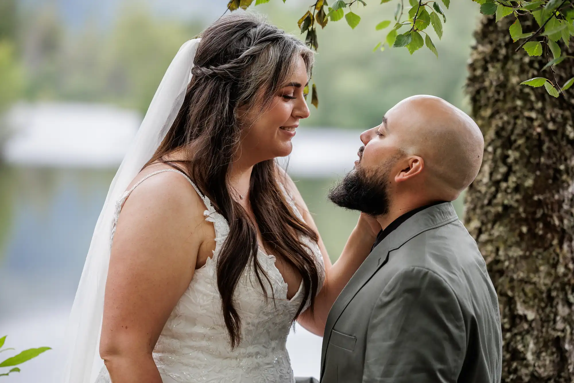 A bride in a white lace wedding dress and long veil gently touches the chin of a bald groom with a beard, both smiling warmly at each other outdoors near a tree, with greenery and water blurred in the background.