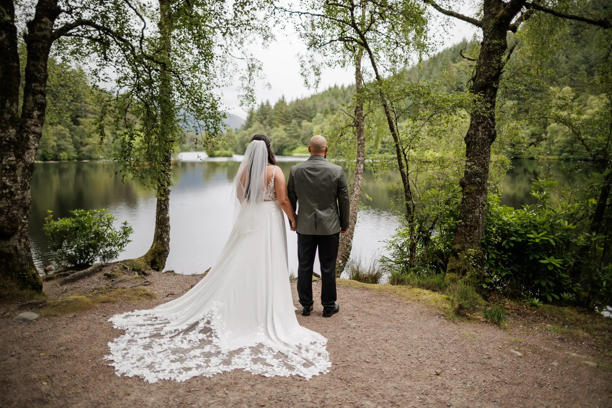 A bride and groom stand hand in hand, facing a calm lake surrounded by lush green trees. The bride wears a long white gown with a veil, and the groom is in a dark suit. They are viewed from behind, framed by tall trees and nature.