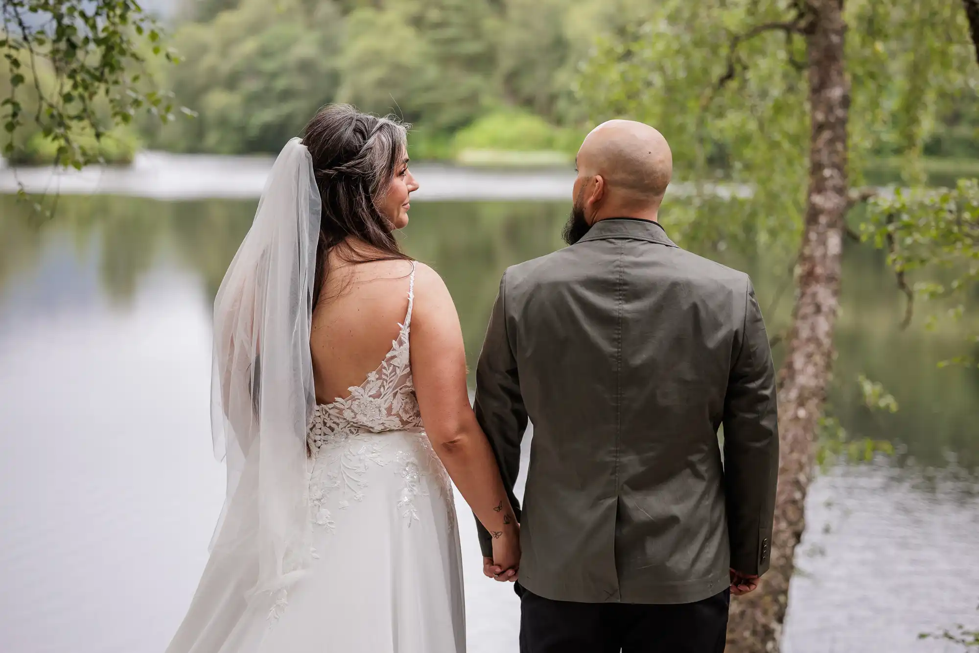 A bride in a white lace dress and veil stands holding hands with a bald, bearded groom in a grey jacket, both seen from behind, by a serene lake surrounded by green trees. The bride looks at the groom and smiles.