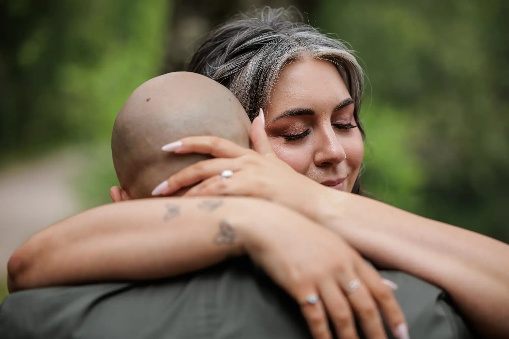 A woman with long, grey-streaked hair hugs a bald person. Her eyes are closed and she looks peaceful. Her hands, with rings and small tattoos, rest gently on the person's neck. The background is a blurred green outdoor scene.