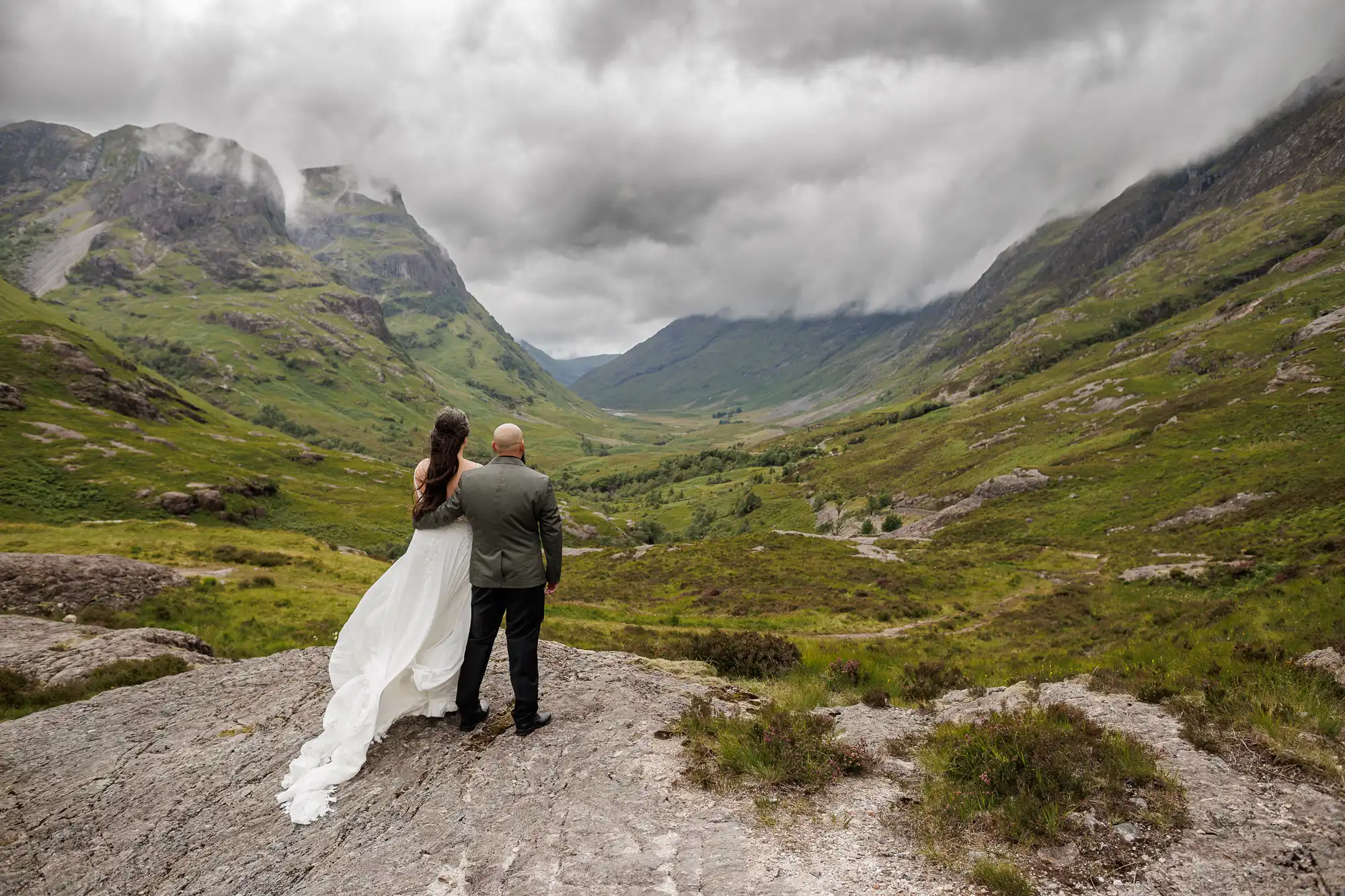 A bride and groom stand on a rocky ledge, backs to the camera, overlooking a lush green valley surrounded by dramatic mountains under a cloudy sky. The bride's white dress flows as they gaze at the scenic, misty landscape ahead.
