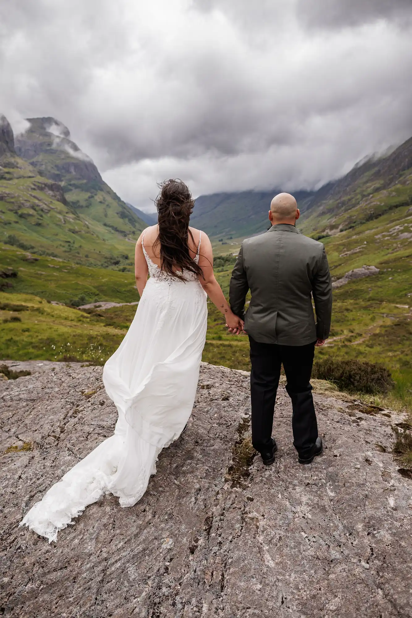 A couple, seen from behind, stands on a rocky ledge in a mountainous, green landscape. The woman wears a flowing white dress, and the man wears a dark suit. They hold hands and face dramatic, cloudy skies and distant, misty peaks.