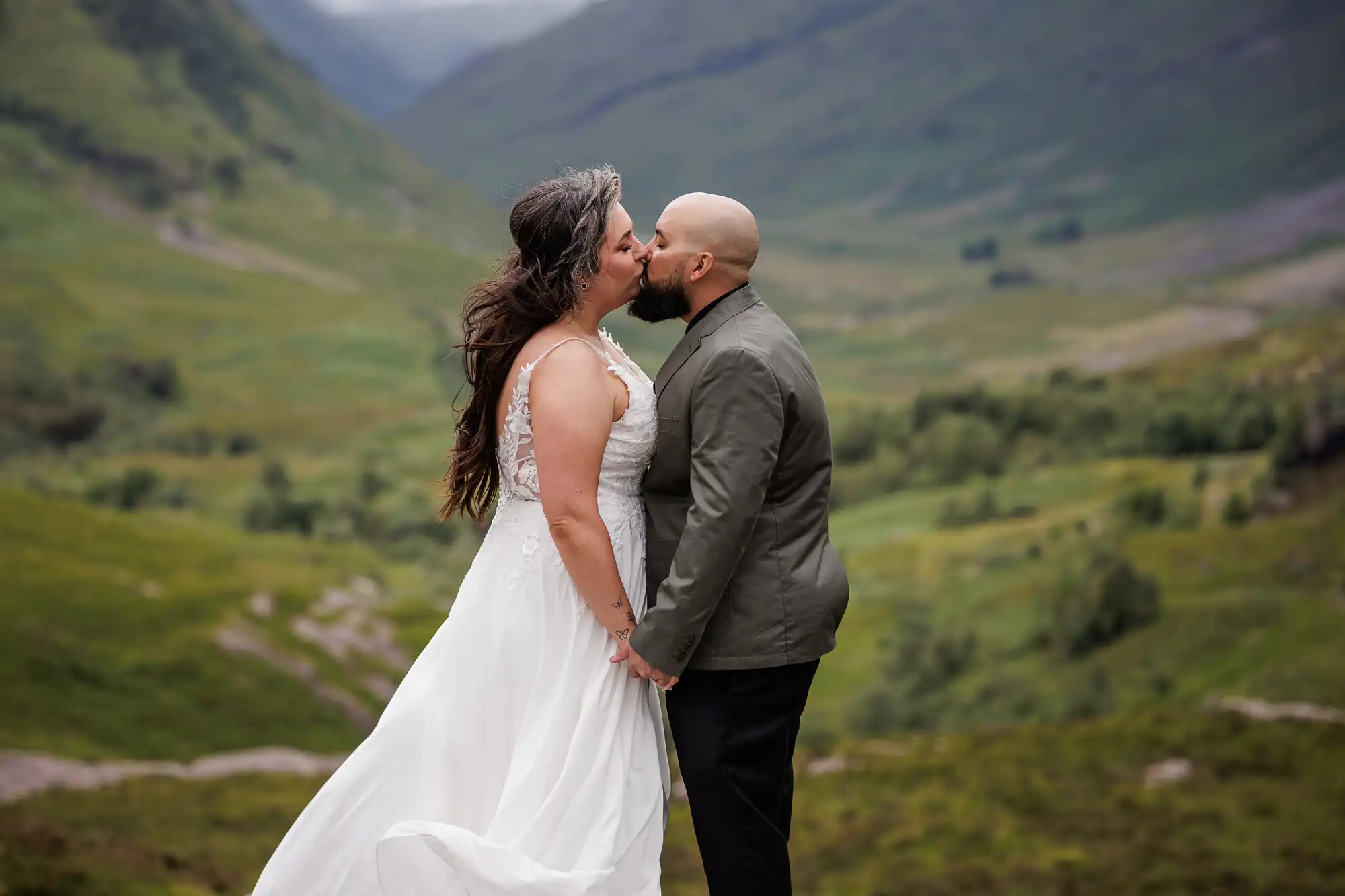 A couple stands outdoors on a lush green hillside, holding hands and sharing a kiss. The woman wears a flowing white dress, and the man wears a green jacket and dark trousers. Rolling hills and misty mountains form a scenic, natural background.