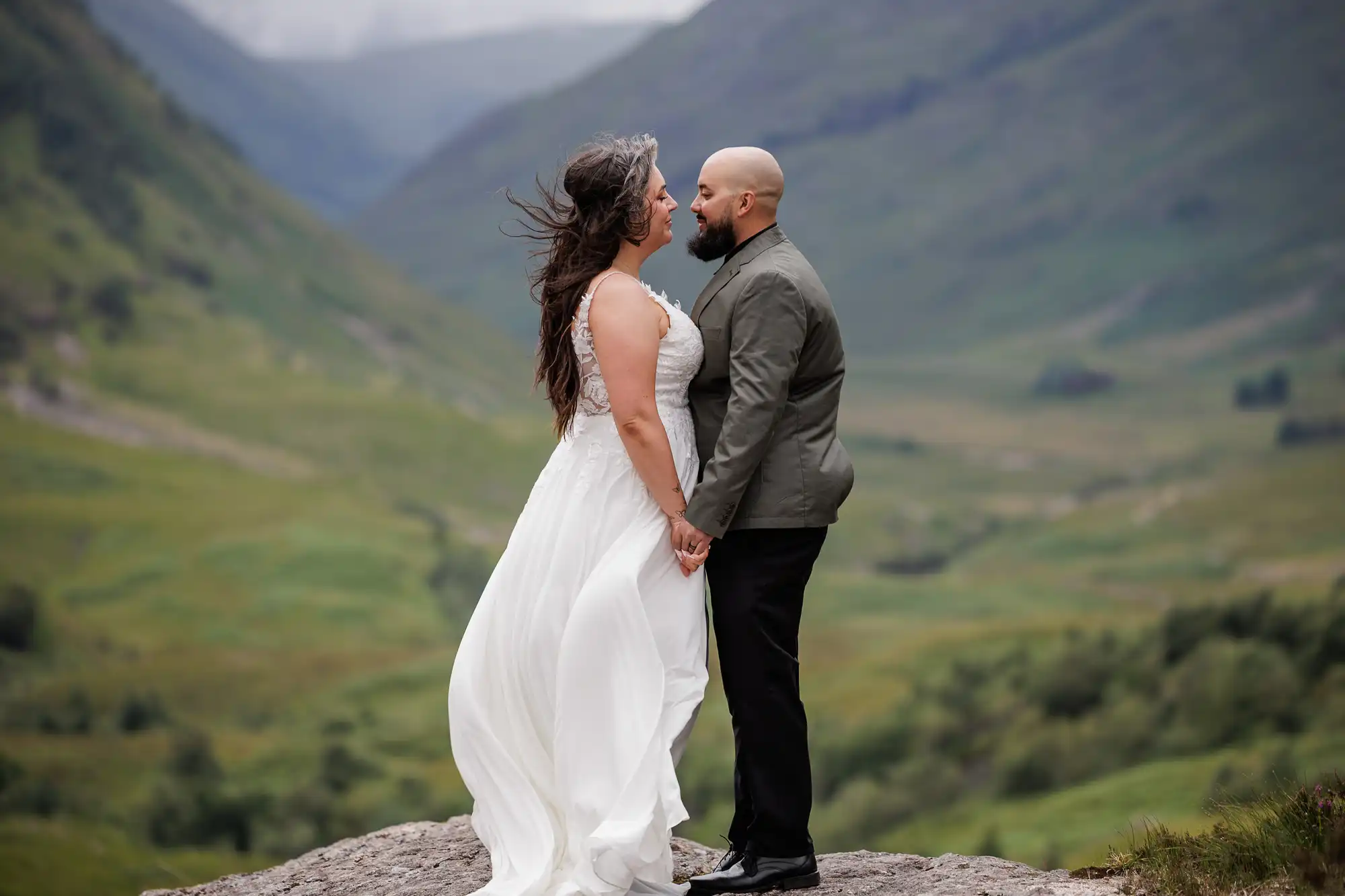 A couple stands on a rock in a lush, green valley. The woman, in a flowing white dress, faces a bearded man in a grey jacket and black trousers. They hold hands, gazing at each other, with mountains and a cloudy sky in the background.