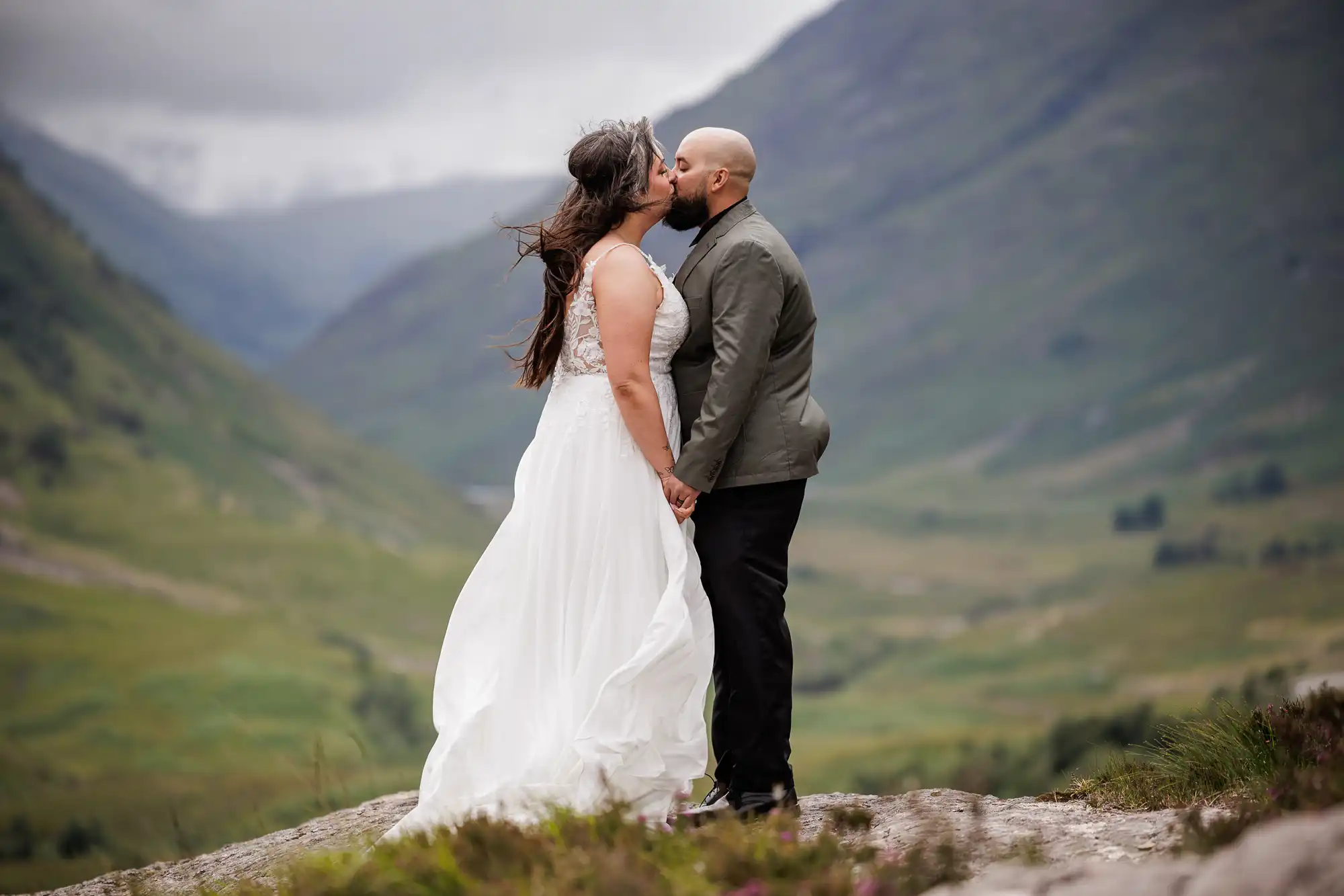Glencoe elopement photographer image of a bride in a flowing white dress and a groom in a grey jacket kiss whilst standing on a rocky hilltop, surrounded by lush green mountains under a cloudy sky. The bride’s hair blows in the wind, and they hold hands, facing each other.
