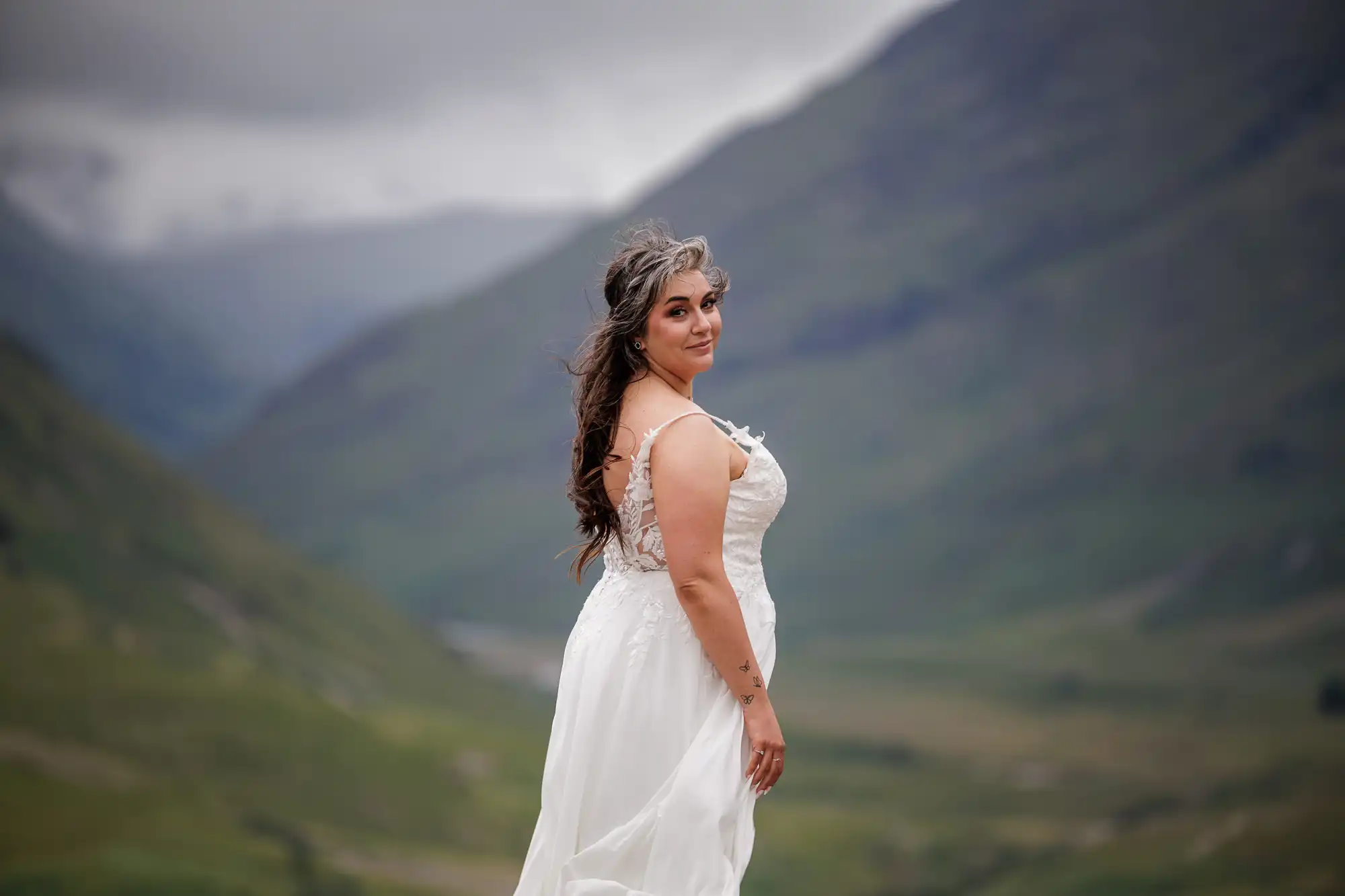 A bride in a flowing white dress stands outdoors, looking over her shoulder. Her long hair is windswept. Lush green mountains and a cloudy sky form a dramatic natural backdrop, creating a serene and picturesque scene.