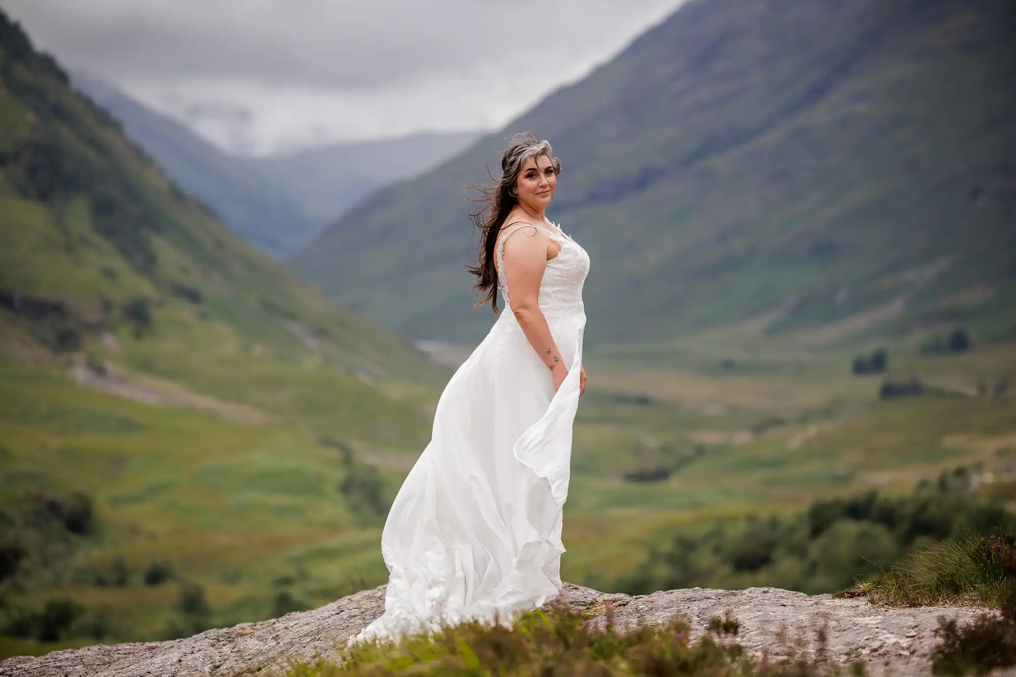 A woman in a flowing white wedding dress stands on a rocky hilltop, smiling slightly, with lush green mountains and a cloudy sky in the background. Her long brown hair blows gently in the wind, and she is turned slightly to the side.