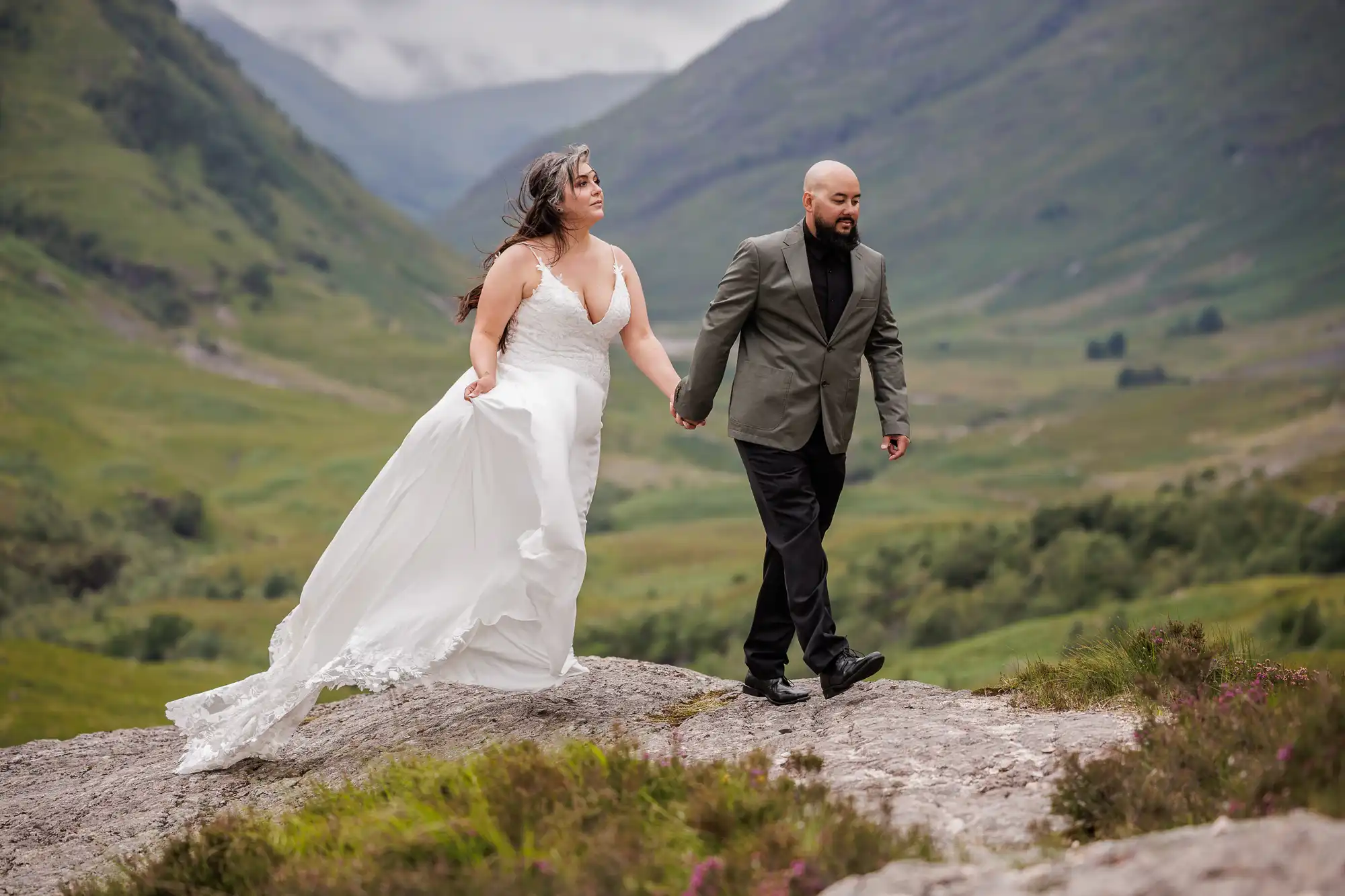 A bride in a flowing white dress and a groom in a grey jacket with black trousers walk hand in hand on a rocky ledge in a lush green mountain valley, with misty hills and dramatic clouds in the background.