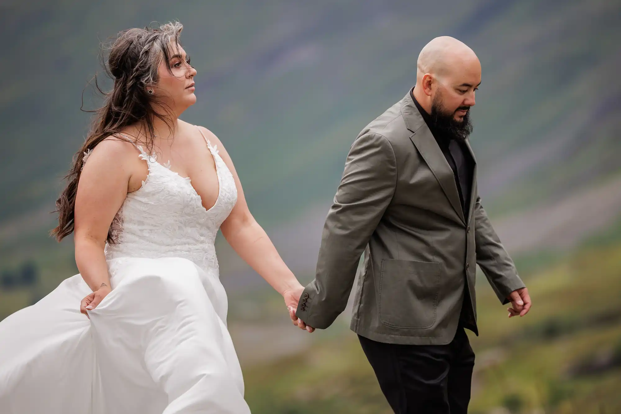 A bride in a white lace wedding dress holds hands with a groom in a dark suit. They walk outdoors with green hills in the background. The bride’s hair is windswept, and both have serious expressions, creating a candid, intimate moment.