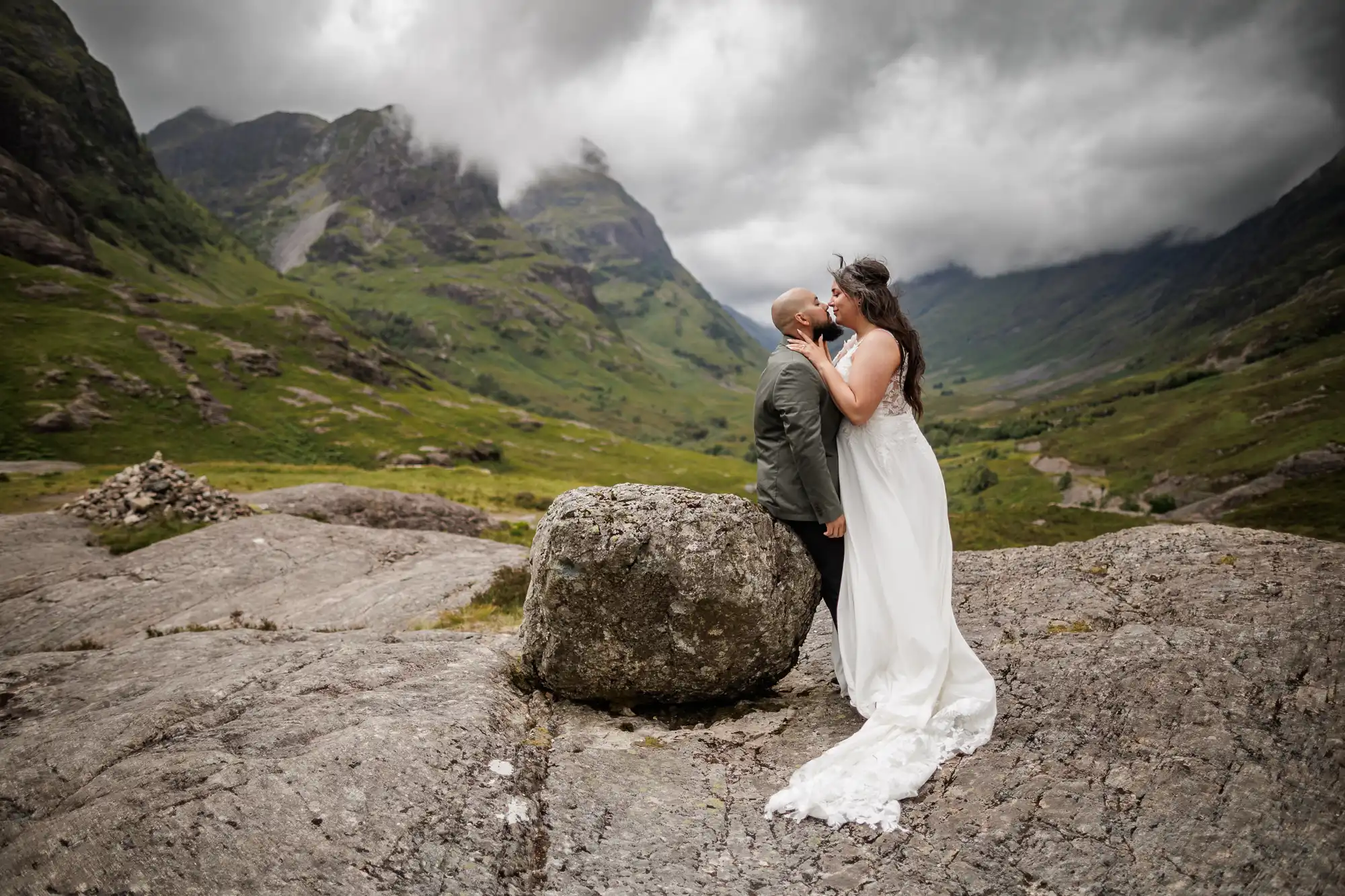 A bride in a long white dress kisses a person in a green suit on a rocky hill. Misty, green mountains and dramatic clouds fill the background, creating a romantic and wild landscape. The bride stands on a rock, making her taller than her partner.