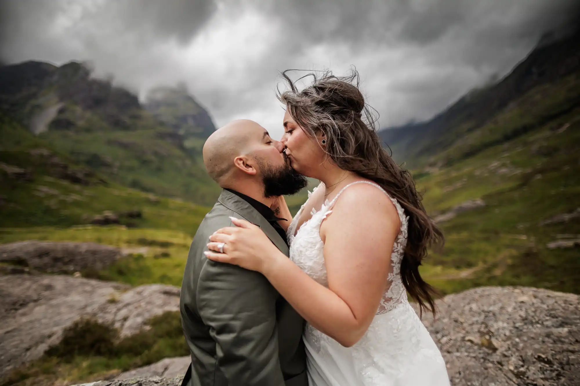 A couple in wedding attire kisses outdoors on rocky terrain, surrounded by green hills and dramatic, cloudy skies. The woman wears a white lace dress; the man wears a dark suit. Wind blows her hair as they embrace intimately among the scenic landscape.