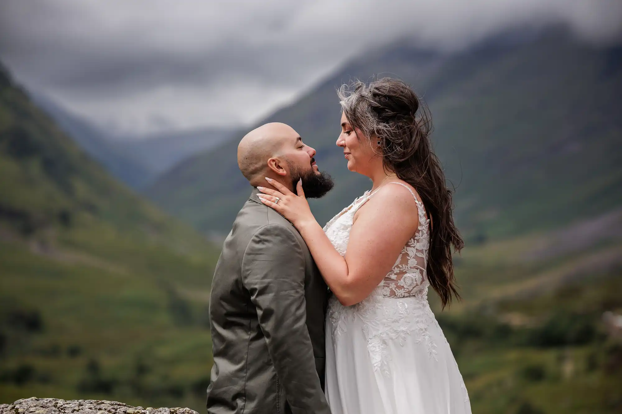 A couple stand close together outdoors, facing each other lovingly. The woman, in a white lace wedding dress, holds the man’s face. The man, wearing a suit jacket, gazes up at her. Behind them are green mountains and a cloudy sky.