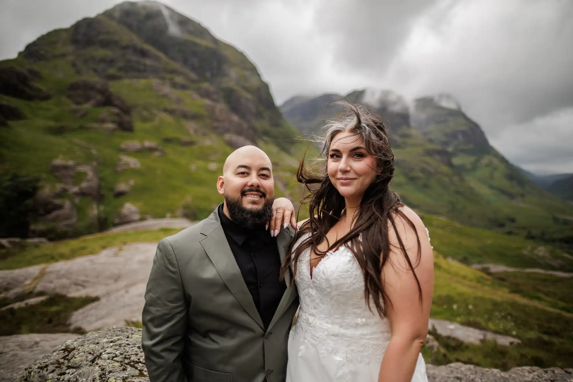 A couple pose outdoors in front of green, rocky hills. The man wears a green suit and black shirt, smiling, while the woman in a white wedding dress smiles with her hair blowing in the wind. The sky is cloudy and the landscape appears lush and mountainous.