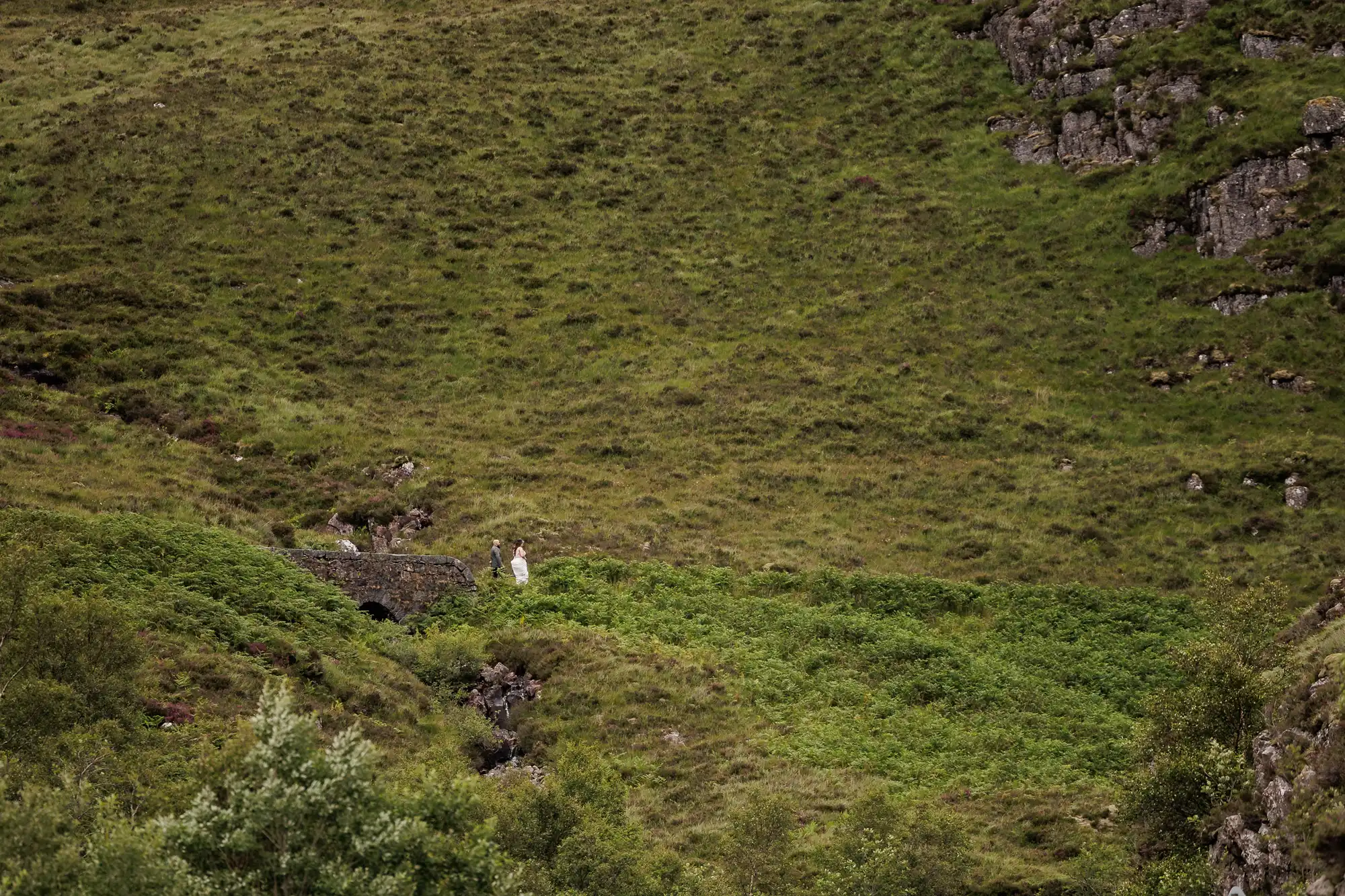 A wide, lush green hillside dotted with rocks and bushes. At the lower centre, two small human figures in light clothing stand near a stone structure, almost blending into the landscape’s natural, earthy tones.