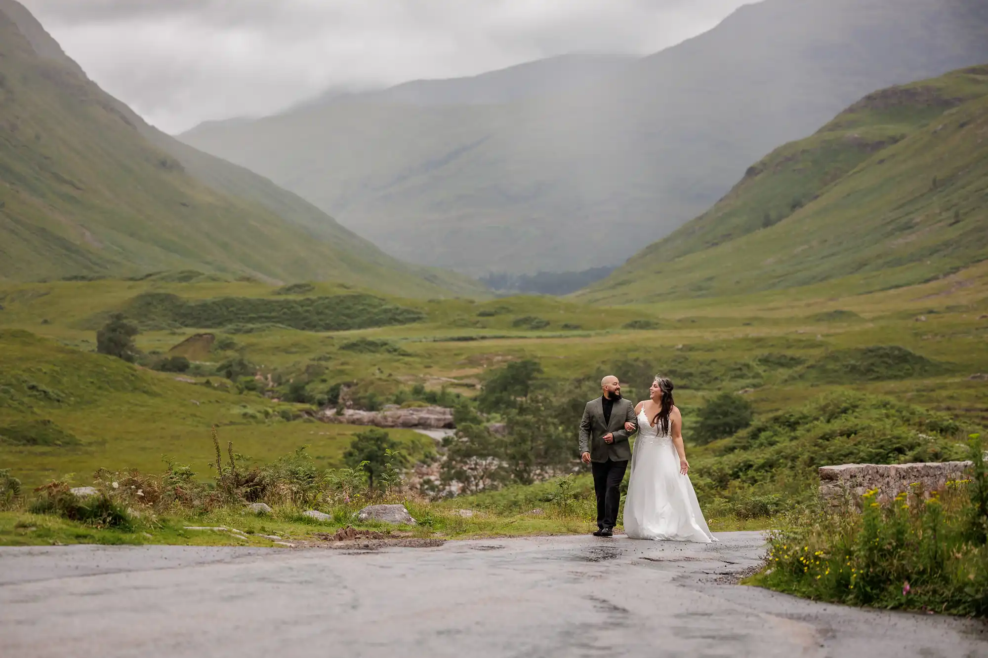 A newlywed couple walk hand-in-hand on a winding road in a lush, green valley surrounded by misty hills. The groom wears a dark suit and the bride wears a white gown with a flower crown. The scene looks peaceful and romantic under a cloudy sky.