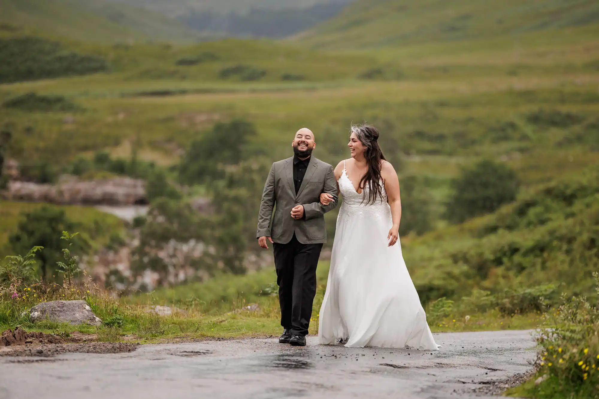 A smiling couple walk arm-in-arm outdoors; the man wears a dark suit, and the woman wears a white wedding dress. They are on a wet road surrounded by lush green hills and grass under an overcast sky, creating a joyful, serene scene.