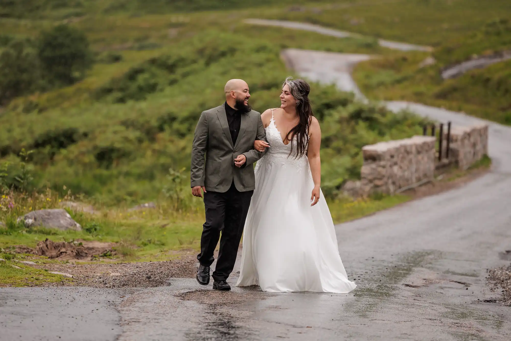 A smiling couple walks arm-in-arm down a wet country road. The person on the left wears a green jacket and black trousers, while the person on the right wears a white wedding dress. Lush greenery and a stone bridge are visible in the background.
