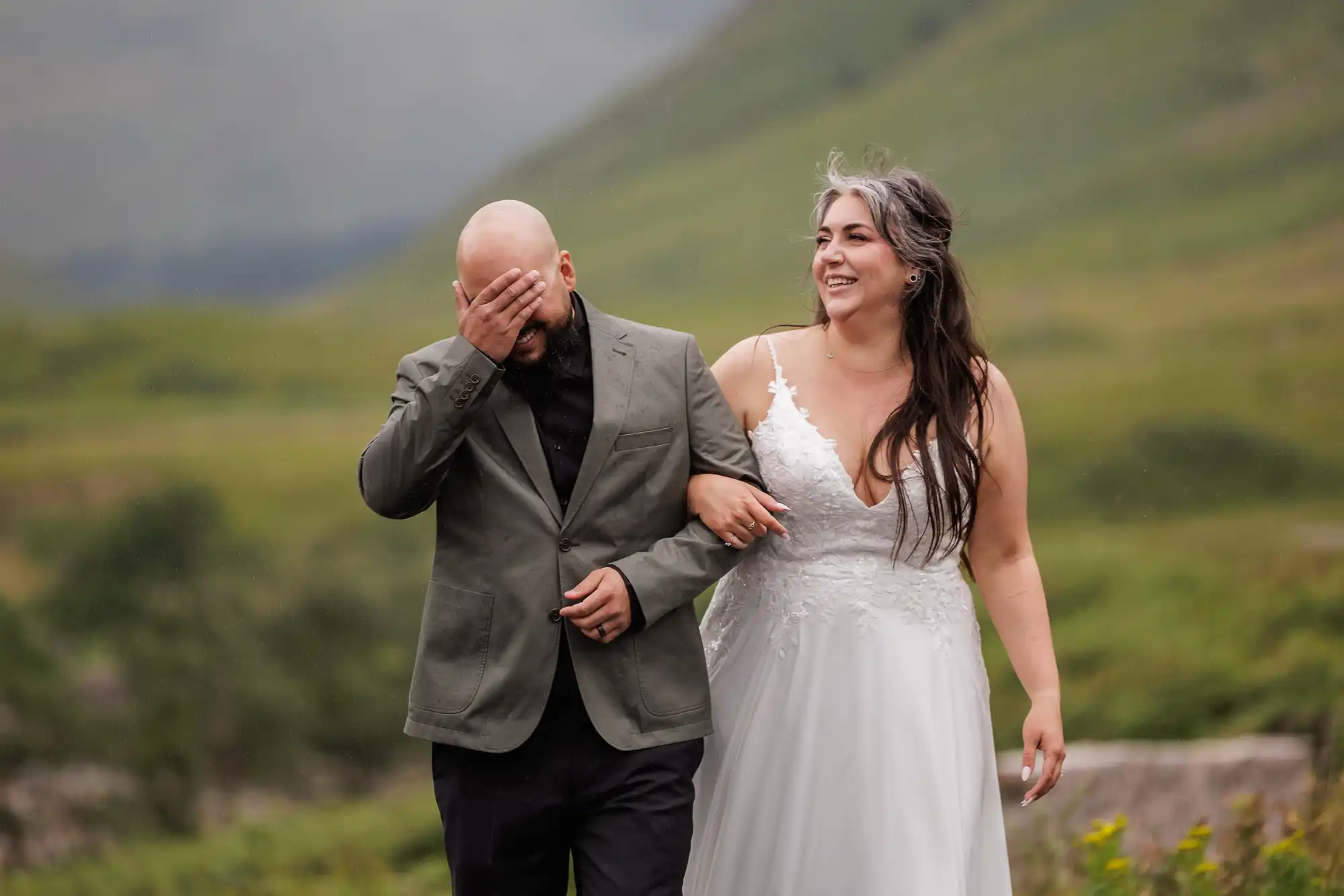 A bride in a white wedding dress smiles, linking arms with a man in a grey blazer and black shirt, who covers his face with his hand. They walk outdoors on green, hilly terrain, sharing a joyful, candid moment together.