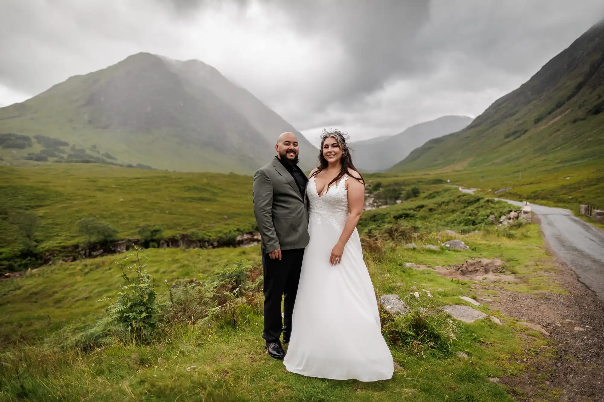 A bride in a white gown and a groom in a suit stand close together on a grassy hillside, smiling. Behind them are misty mountains, green valleys, and a winding road under a cloudy sky, creating a dramatic, scenic backdrop.