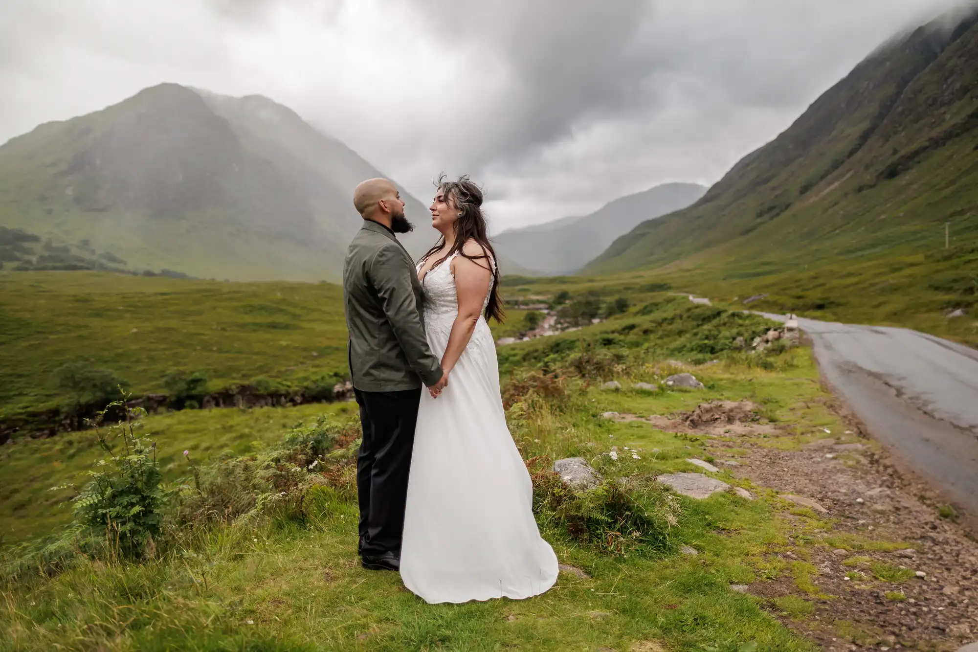 A couple stands holding hands on a grassy hillside by a narrow road, surrounded by misty, green mountains under a cloudy sky. The woman wears a white dress, the man a green jacket and black trousers. Both look at each other, smiling in the dramatic landscape.