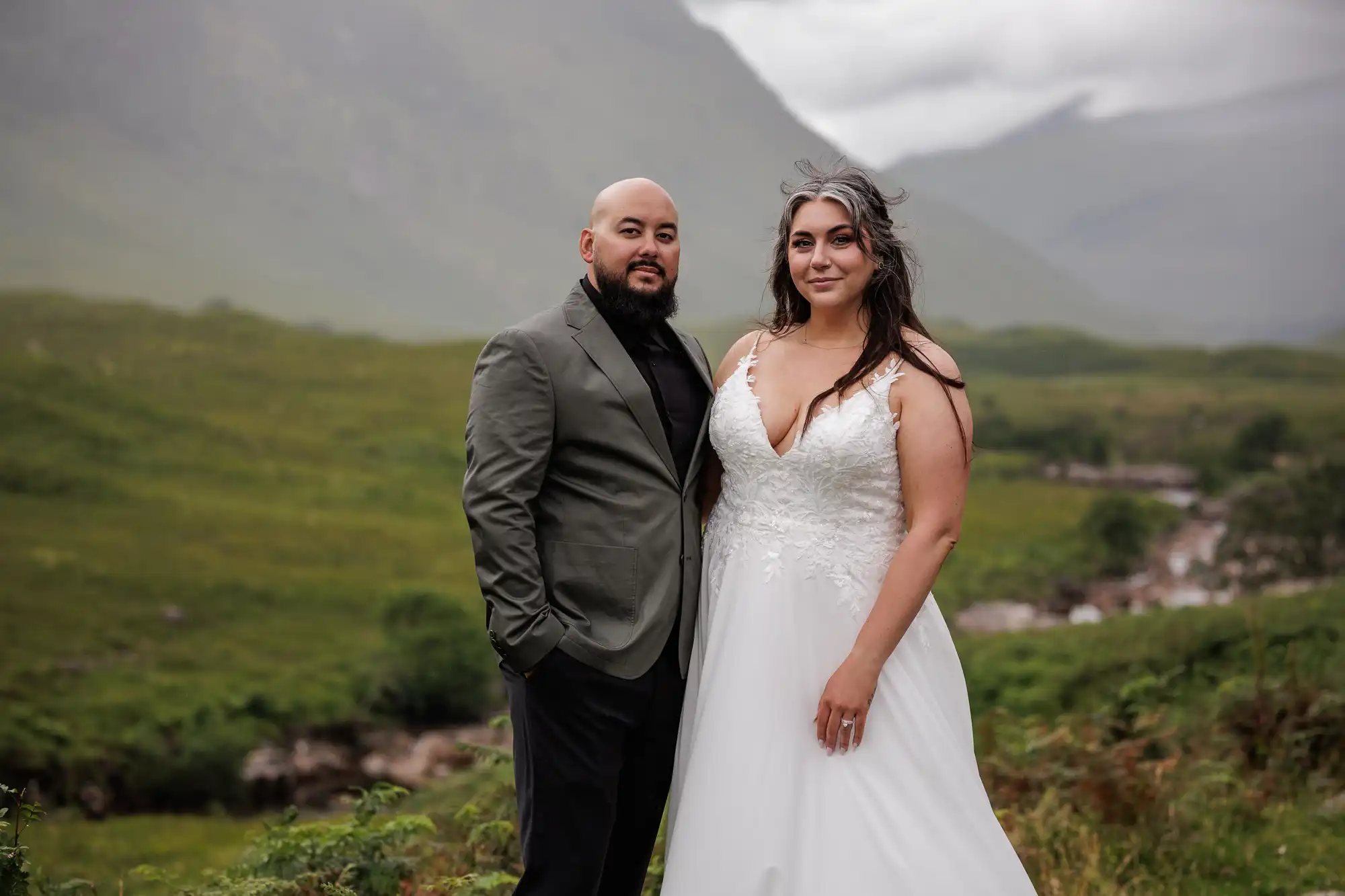 A couple poses outdoors on a green, grassy landscape with mountains and cloudy skies in the background. The person on the left wears a grey jacket and black shirt, while the person on the right wears a white, sleeveless wedding dress with lace detail.
