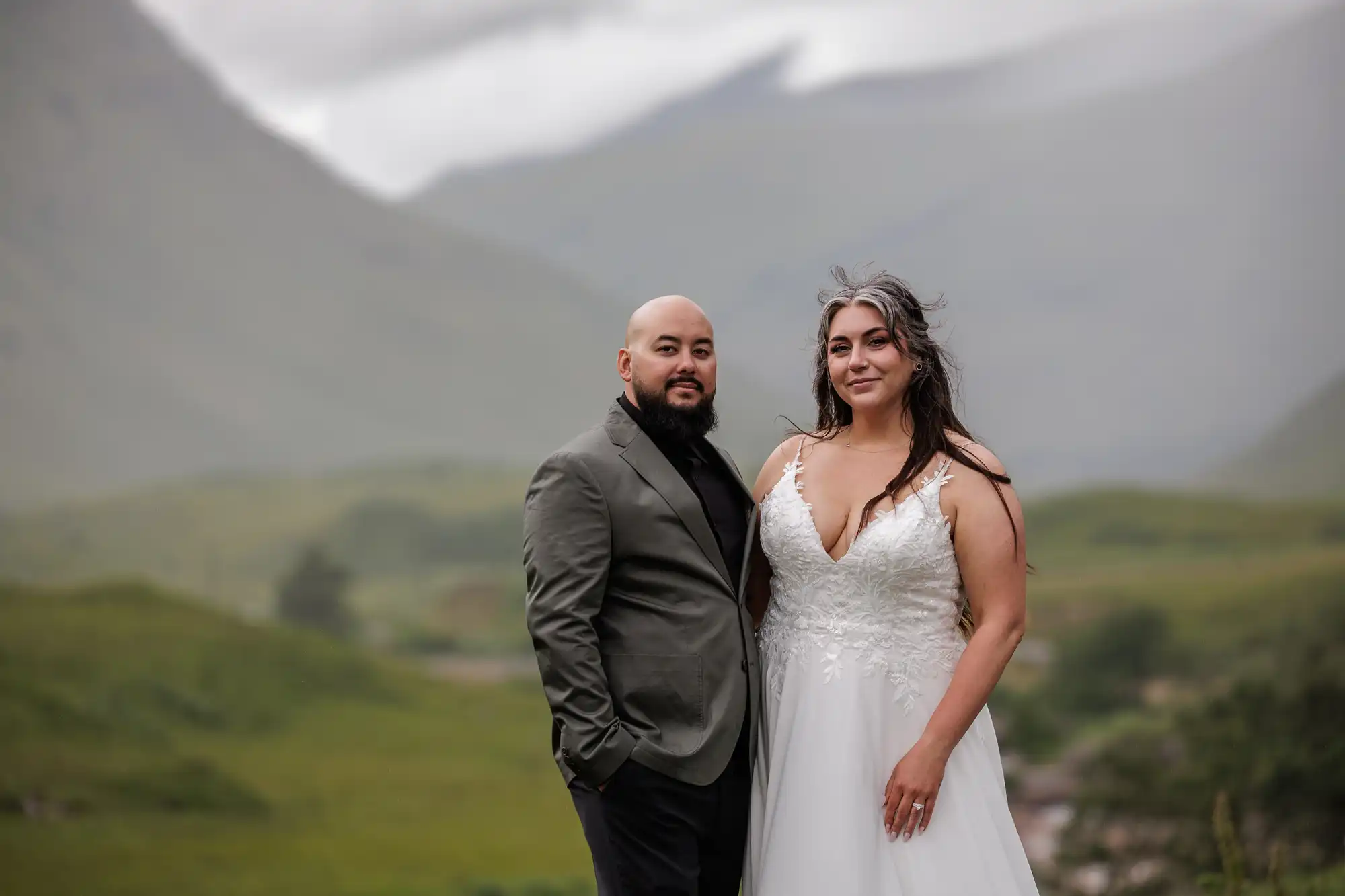 A couple poses outdoors against misty mountains and green hills. The person on the left wears a grey blazer over a black shirt, and the person on the right wears a white wedding dress with lace details and a deep neckline. Both look calmly at the camera.