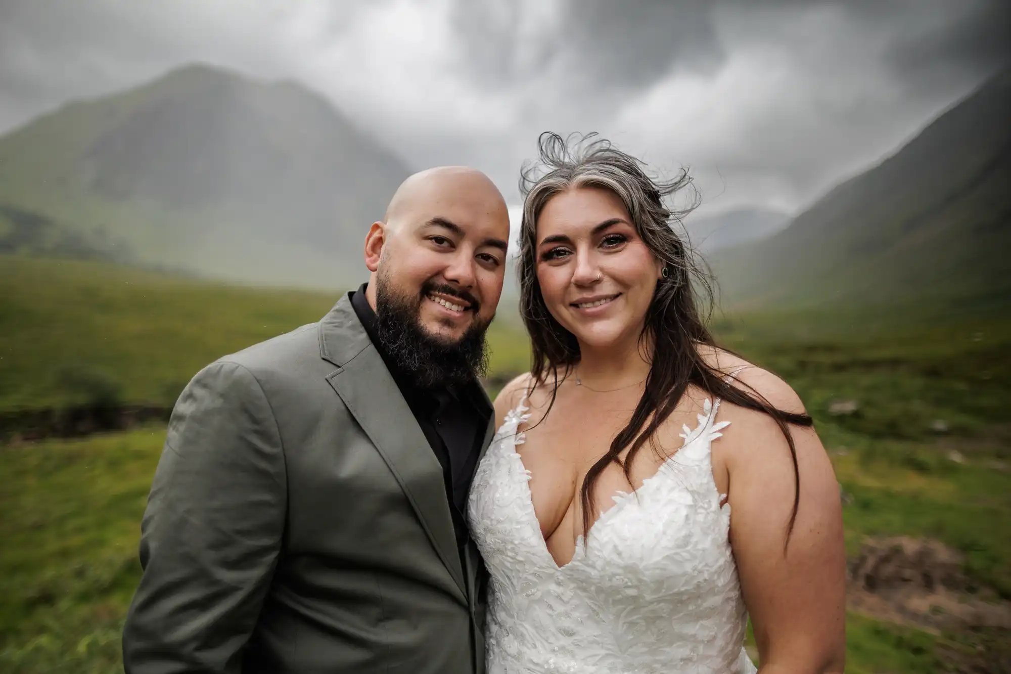 A smiling couple poses outdoors in front of misty green mountains. The man, with a beard and shaven head, wears a grey suit. The woman, with long brown hair and a deep V-neck white lace dress, stands close beside him. The sky is overcast, adding a dramatic effect.
