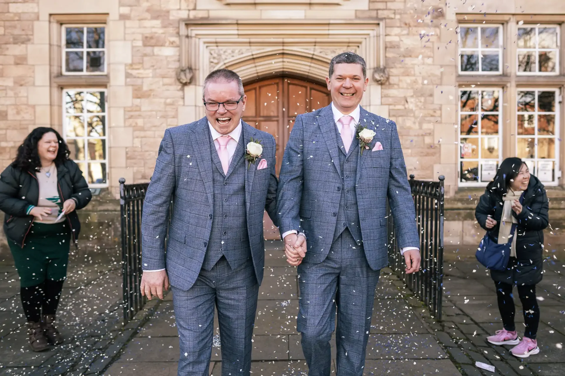 Two newlywed men in matching suits hold hands and smile while walking outside Haddington Registration Office as confetti falls and people stand nearby.