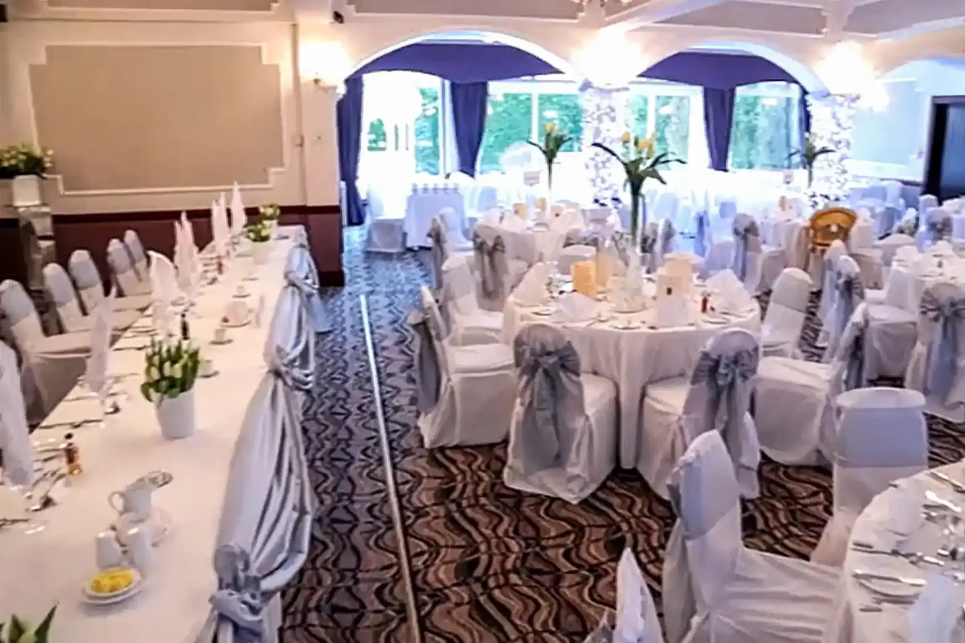 A banquet hall set up for an event at The Hilcroft Hotel, with round and rectangular tables, white chair covers, floral centrepieces, and large windows in the background.