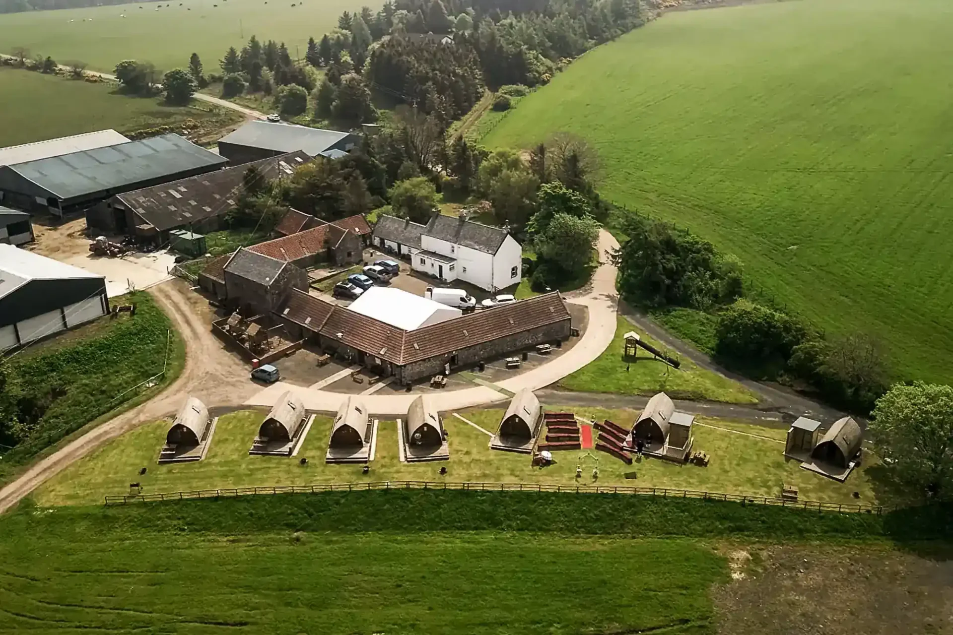 Aerial view of a rural farm, Hilly Cow Wigwams, with several buildings, parked vehicles, and a row of glamping pods on a grassy area surrounded by fields and trees.