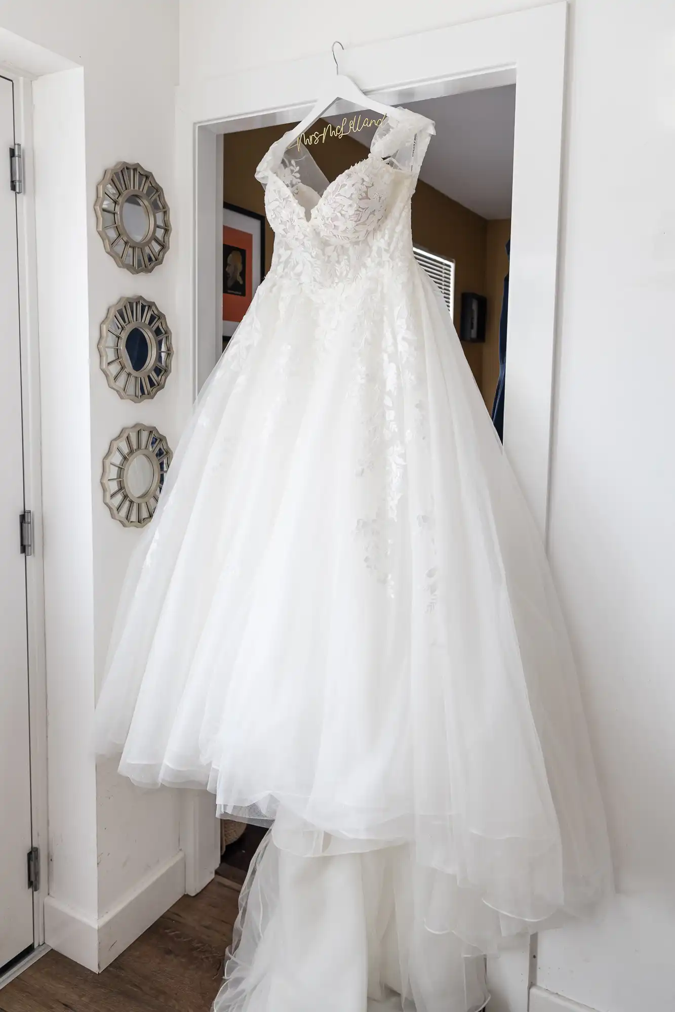 A white lace wedding dress with a full tulle skirt and off-the-shoulder sleeves hangs on a personalised hanger in a doorway. Three decorative mirrors are mounted on the adjacent wall next to the dress.