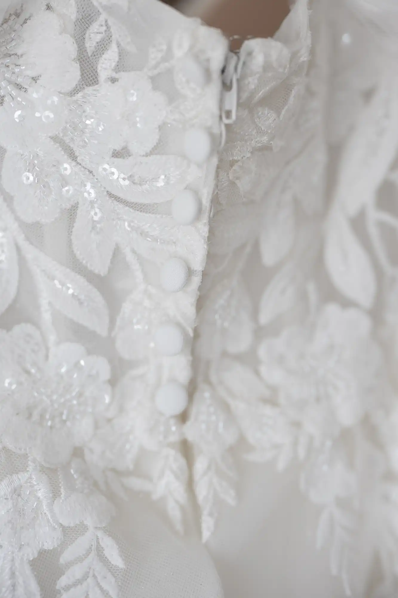 Close-up of an elegant white lace dress with floral embroidery, sequins, and leaf patterns. The dress features a sheer overlay, fabric-covered buttons, and a zip, creating a delicate, intricate, and textured appearance.