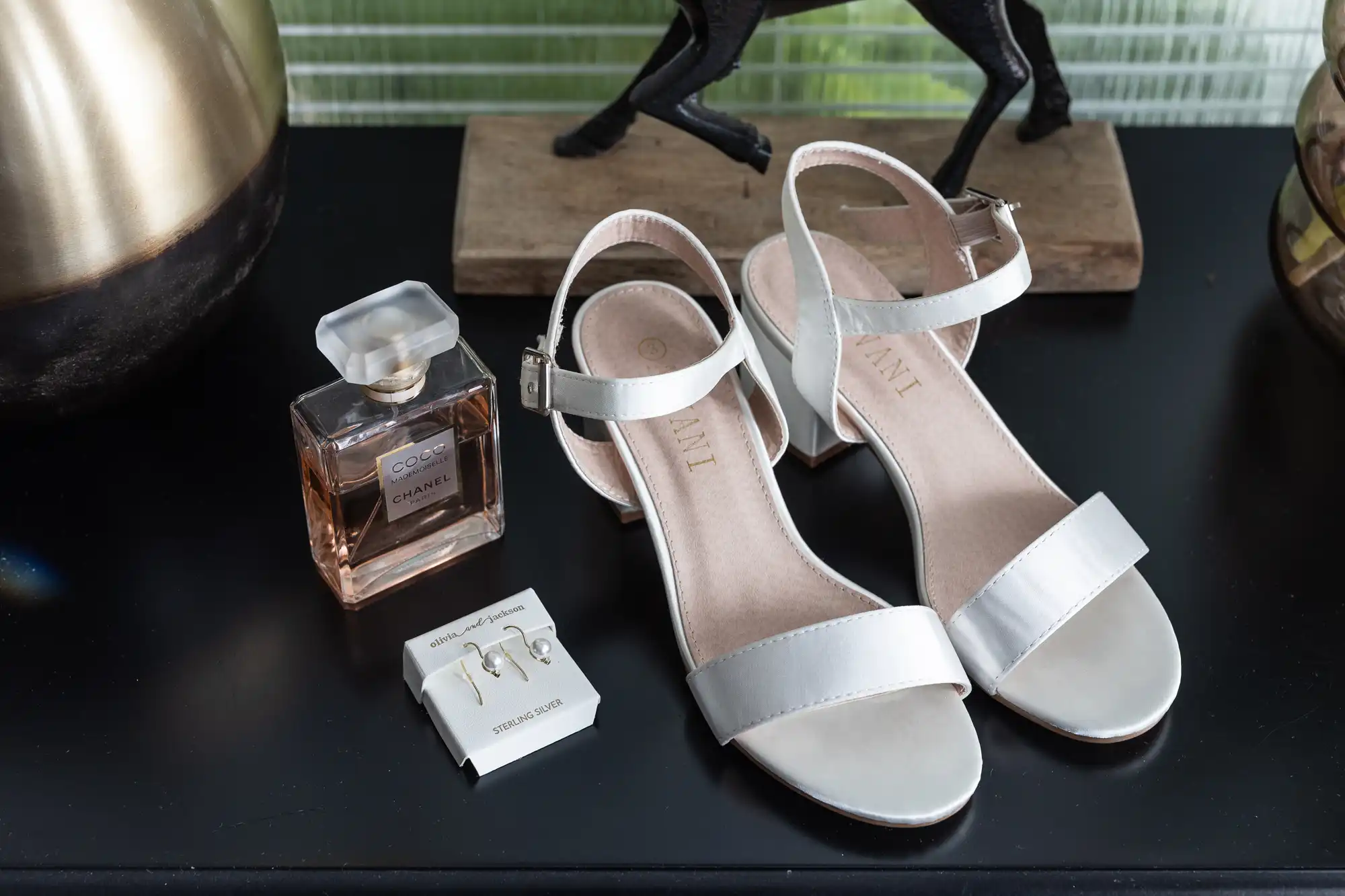 A pair of white, open-toe sandals with ankle straps are displayed on a black surface next to a bottle of Chanel Coco Mademoiselle perfume and a set of silver earrings in a white box, with decorative items in the background.