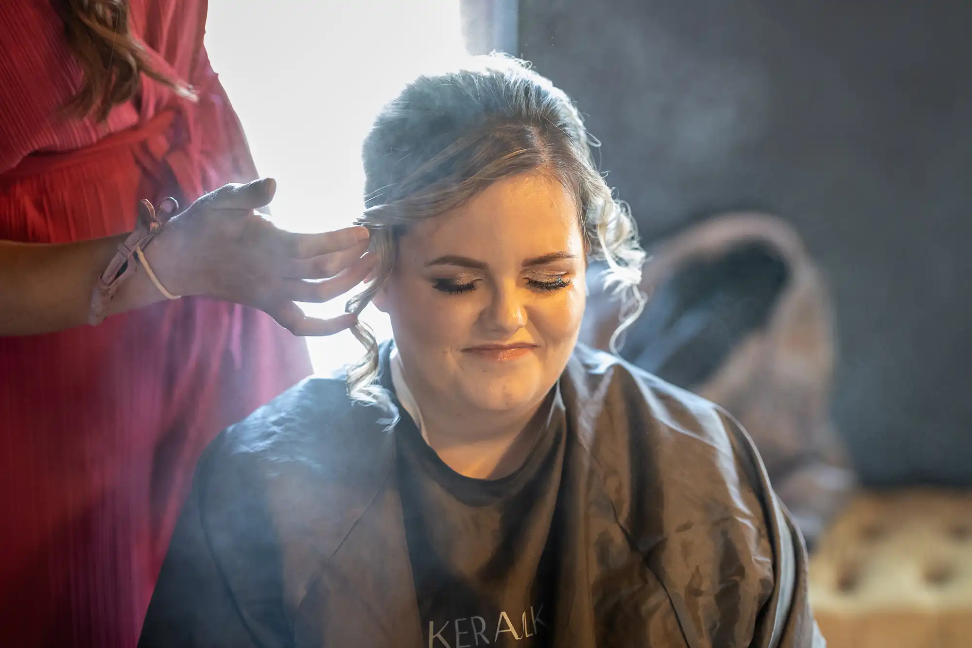 A woman with styled hair and make-up sits with eyes closed, smiling gently. Another person in a red dress is adjusting her hair. Soft light highlights her face, and mist or spray is visible in the air around them, creating a warm, intimate atmosphere.