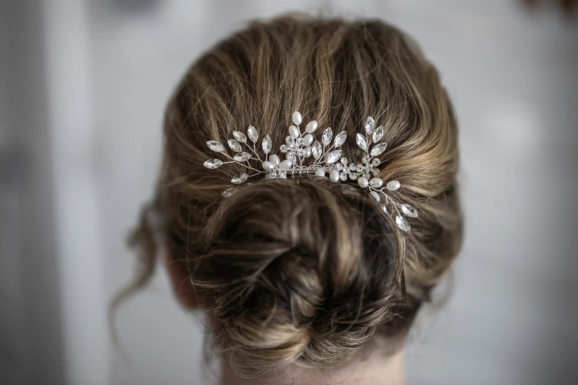 A close-up of the back of a woman’s head shows light brown hair styled in a low, elegant bun adorned with a delicate decorative hairpiece featuring pearls and crystals arranged in a floral and leaf design. The background is softly blurred.