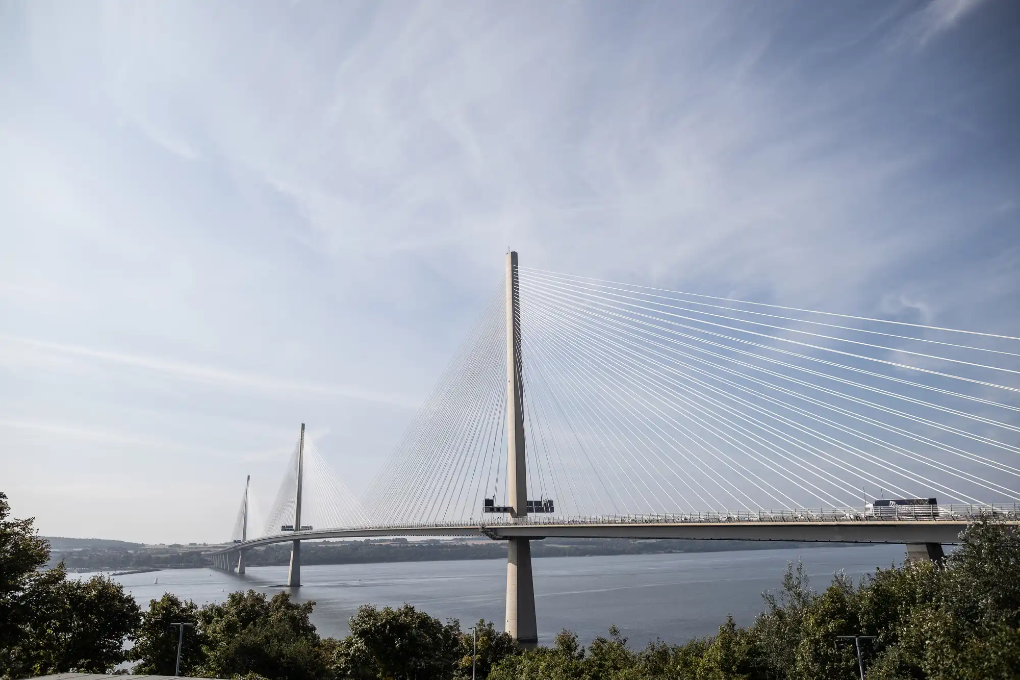 A wide cable-stayed bridge spans across a calm river, supported by tall pylons with numerous white cables. Vehicles cross the bridge, and greenery lines the riverbank below. The sky above is clear with wispy clouds.