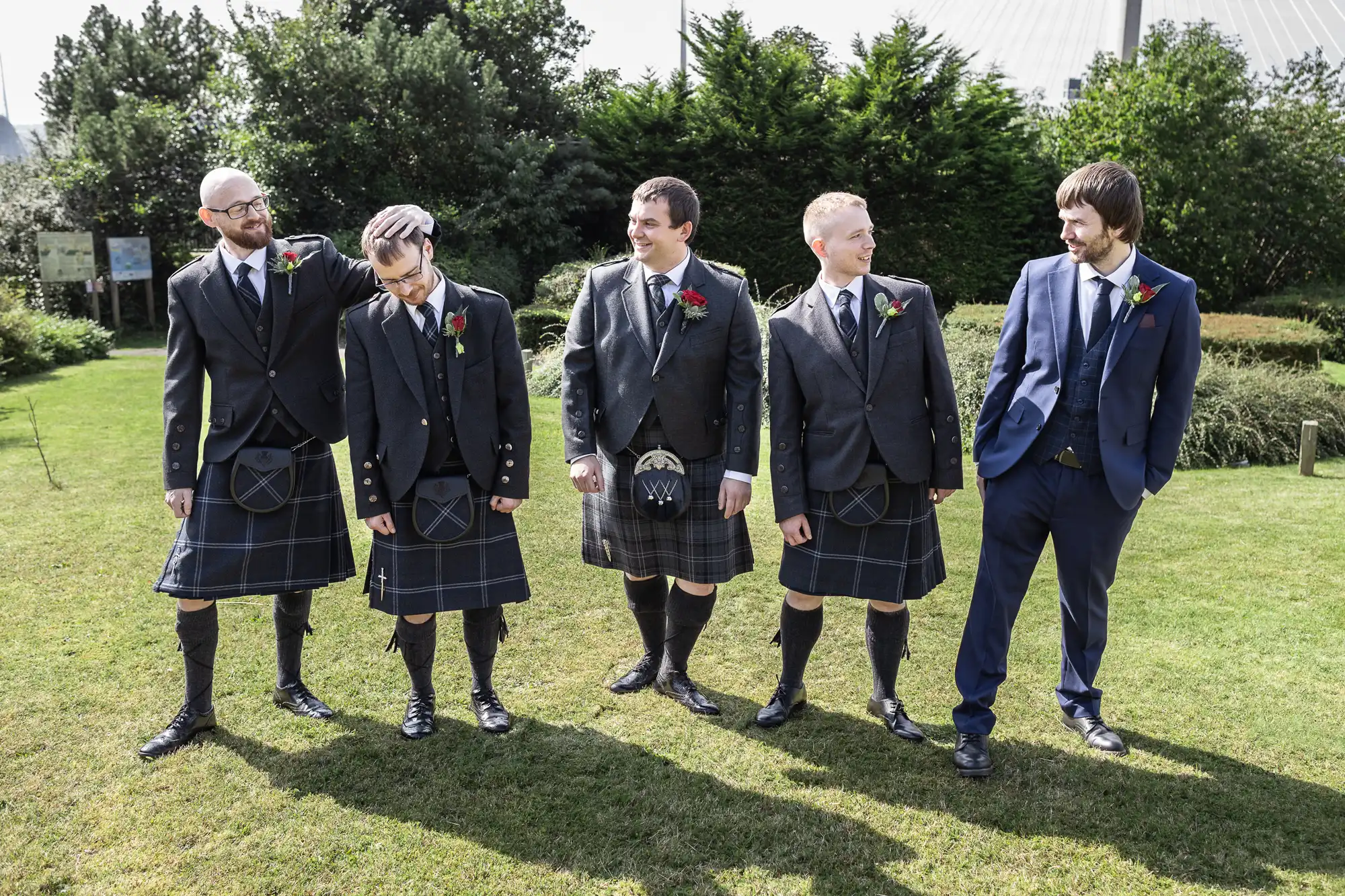 Five men stand on grass in sunlight, four wearing dark formal jackets, shirts, ties, and kilts with sporrans, and one wearing a blue suit. Three have red buttonholes. They smile and look at each other, with trees and bushes in the background.