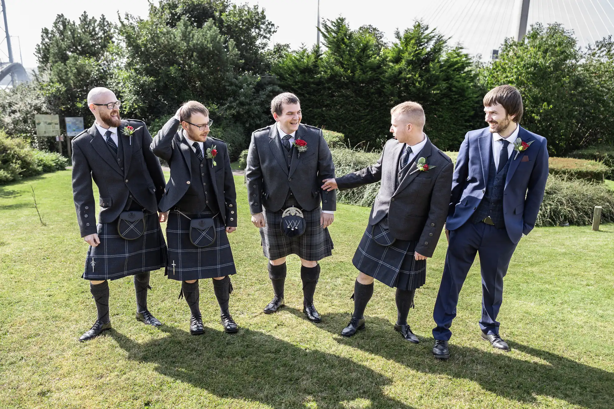 Five men stand together on grass outdoors. Four wear black jackets, waistcoats, kilts, knee-high socks, and buttonholes; one wears a blue suit. They smile and interact cheerfully, with greenery and sunlight in the background.