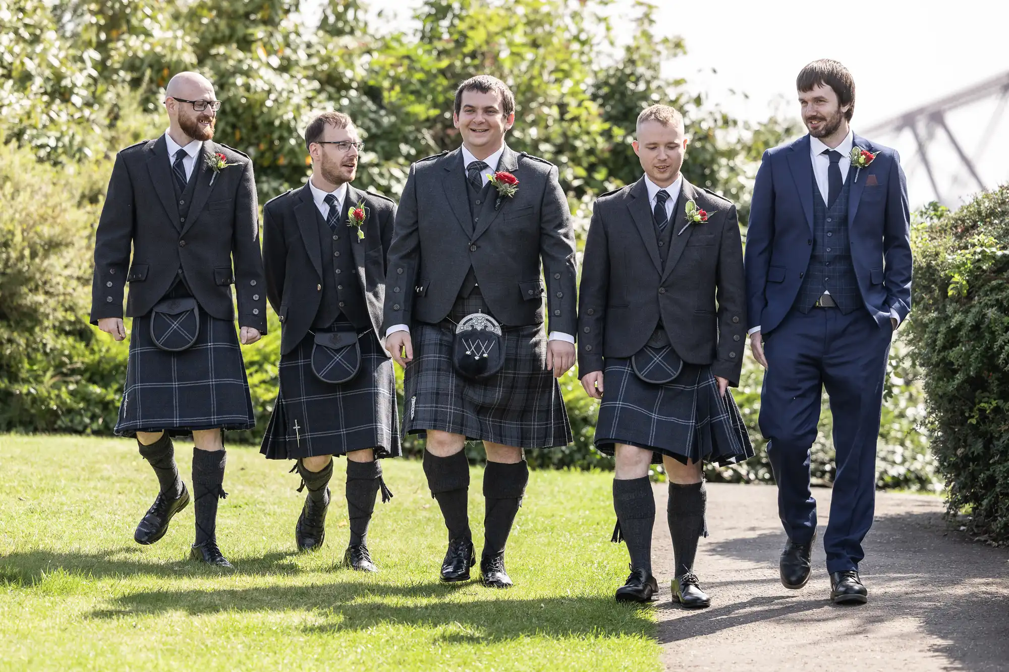 Five men walk outdoors on grass; four wear matching black jackets, ties, and tartan kilts with sporrans, while one wears a blue suit. All have buttonholes. They smile and talk together, with greenery and a bridge visible in the background.