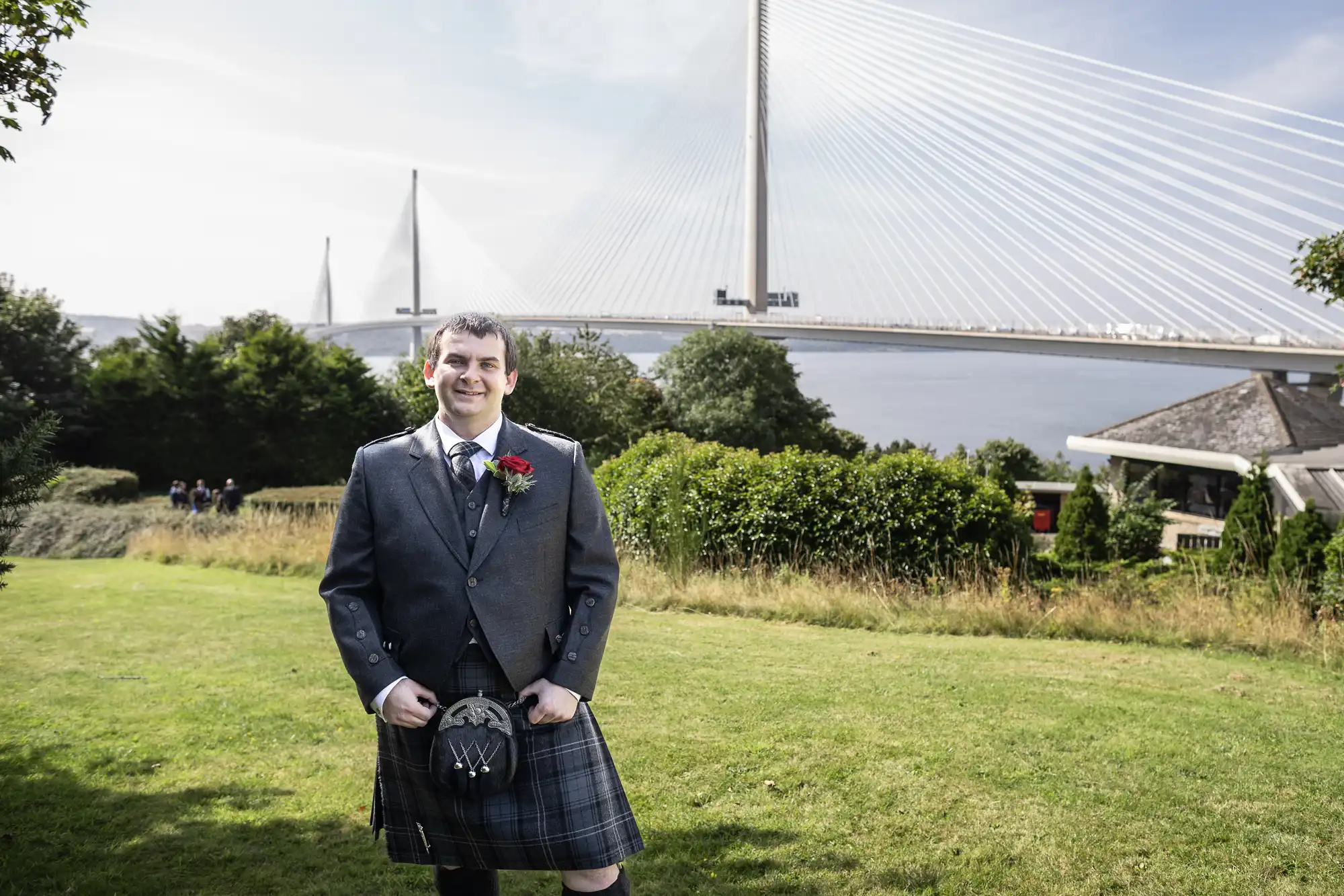A man in a grey jacket and traditional Scottish kilt stands smiling on a grassy lawn. Behind him are green bushes, trees, and a large cable-stayed bridge crossing over water, under a bright, partly cloudy sky.