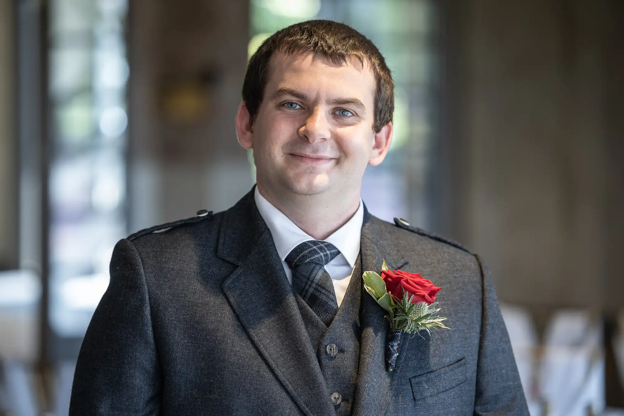 A man with short brown hair and blue eyes smiles at the camera. He wears a dark suit with a checked tie and a red rose buttonhole. The background is softly blurred, showing windows and indoor light.