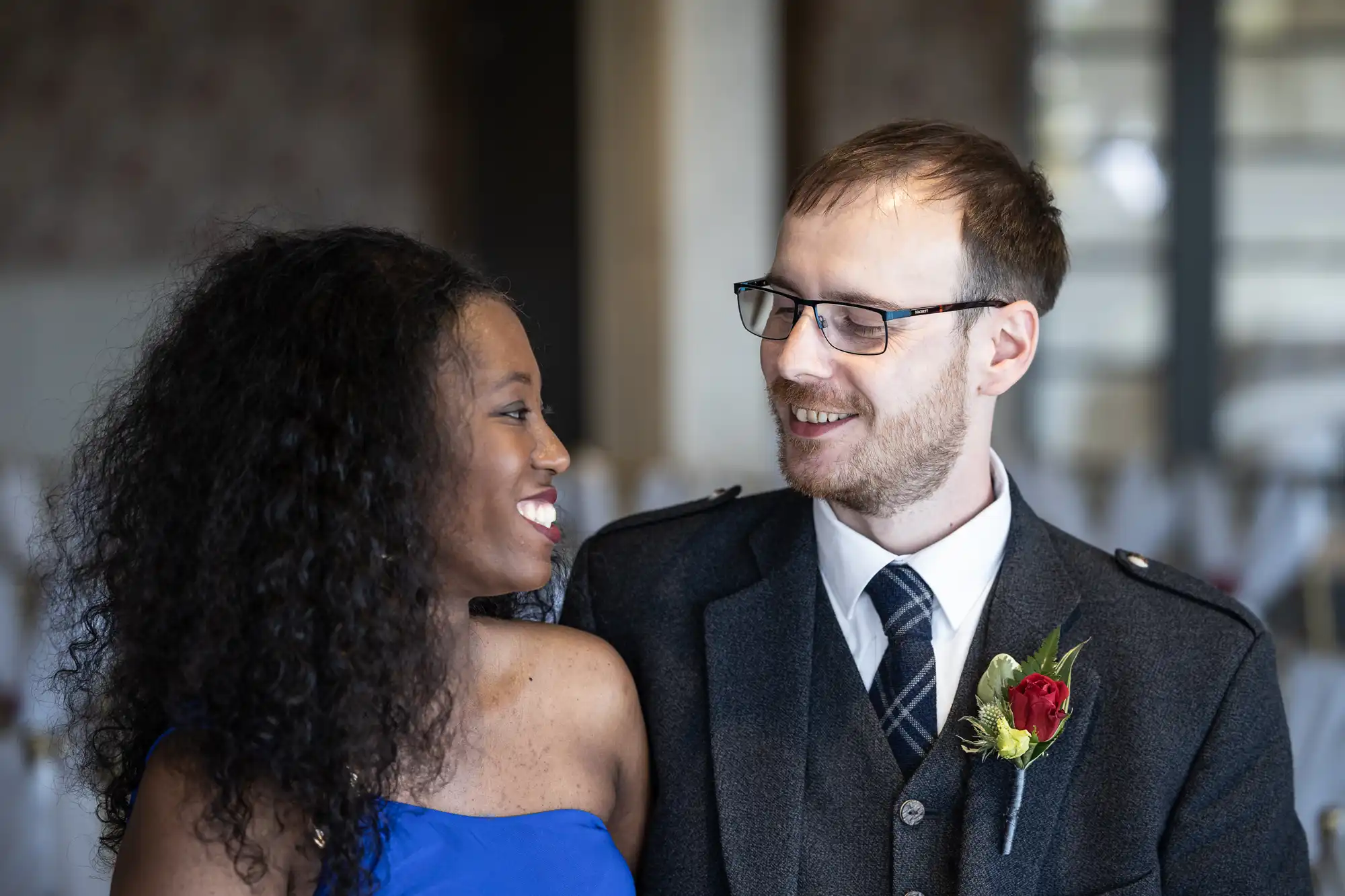 A smiling couple stand close together indoors. The woman has long curly hair and wears a blue one-shoulder dress. The man, in glasses and a dark suit with a checked tie and buttonhole, looks at her warmly. White chairs are blurred in the background.