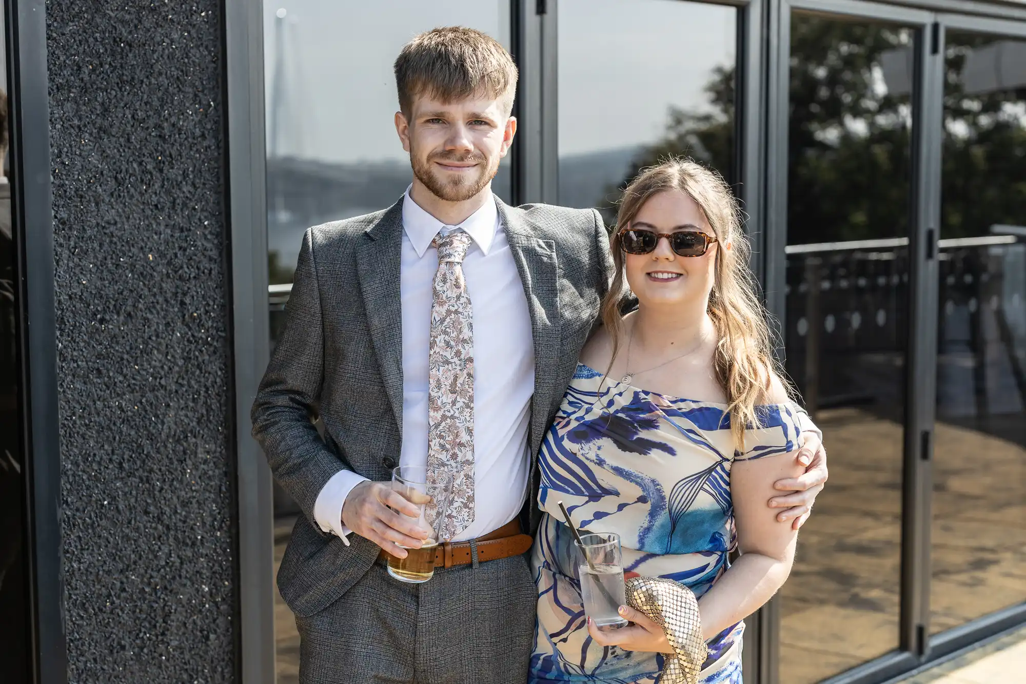 A man in a grey suit with a patterned tie stands beside a woman in a blue and white patterned dress and sunglasses. Both are smiling, holding drinks, and standing outdoors in front of large glass windows on a sunny day.