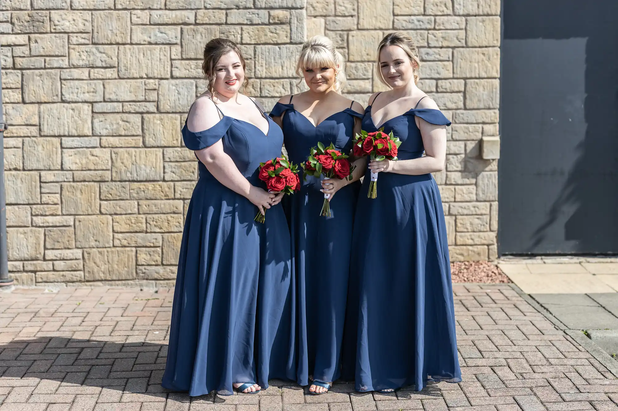 Three women in matching navy blue off-shoulder dresses stand side by side outdoors on a brick path, each holding a bouquet of red and white flowers. They are smiling, and a stone wall with a black door is in the background.