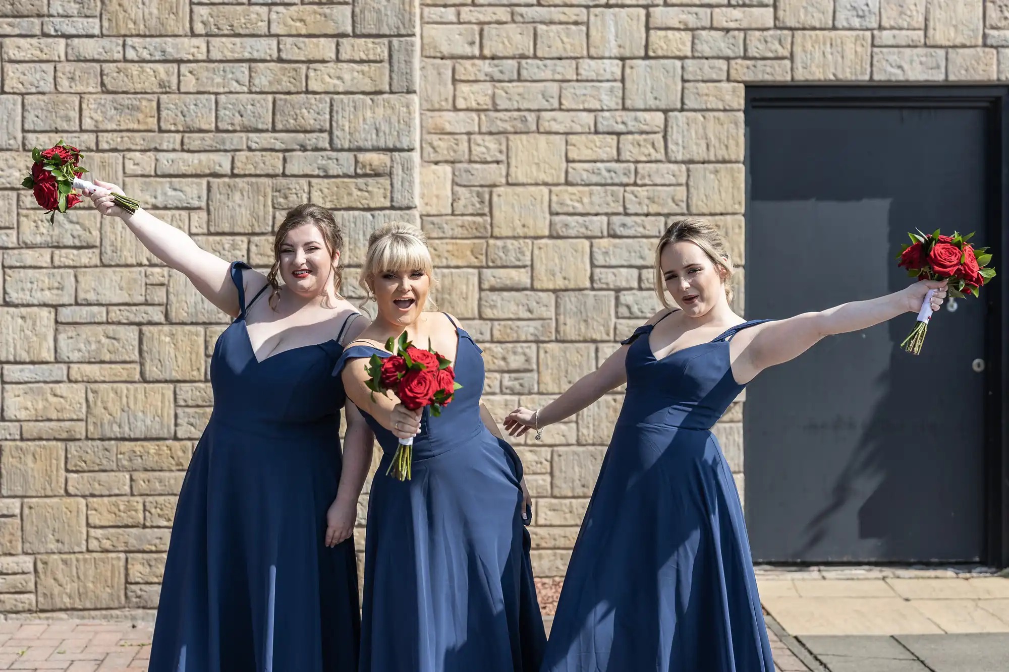 Three women in matching navy blue dresses pose joyfully in front of a stone wall, each holding red rose bouquets. They smile and gesture energetically, with arms outstretched, standing together on a sunny day near a dark door.