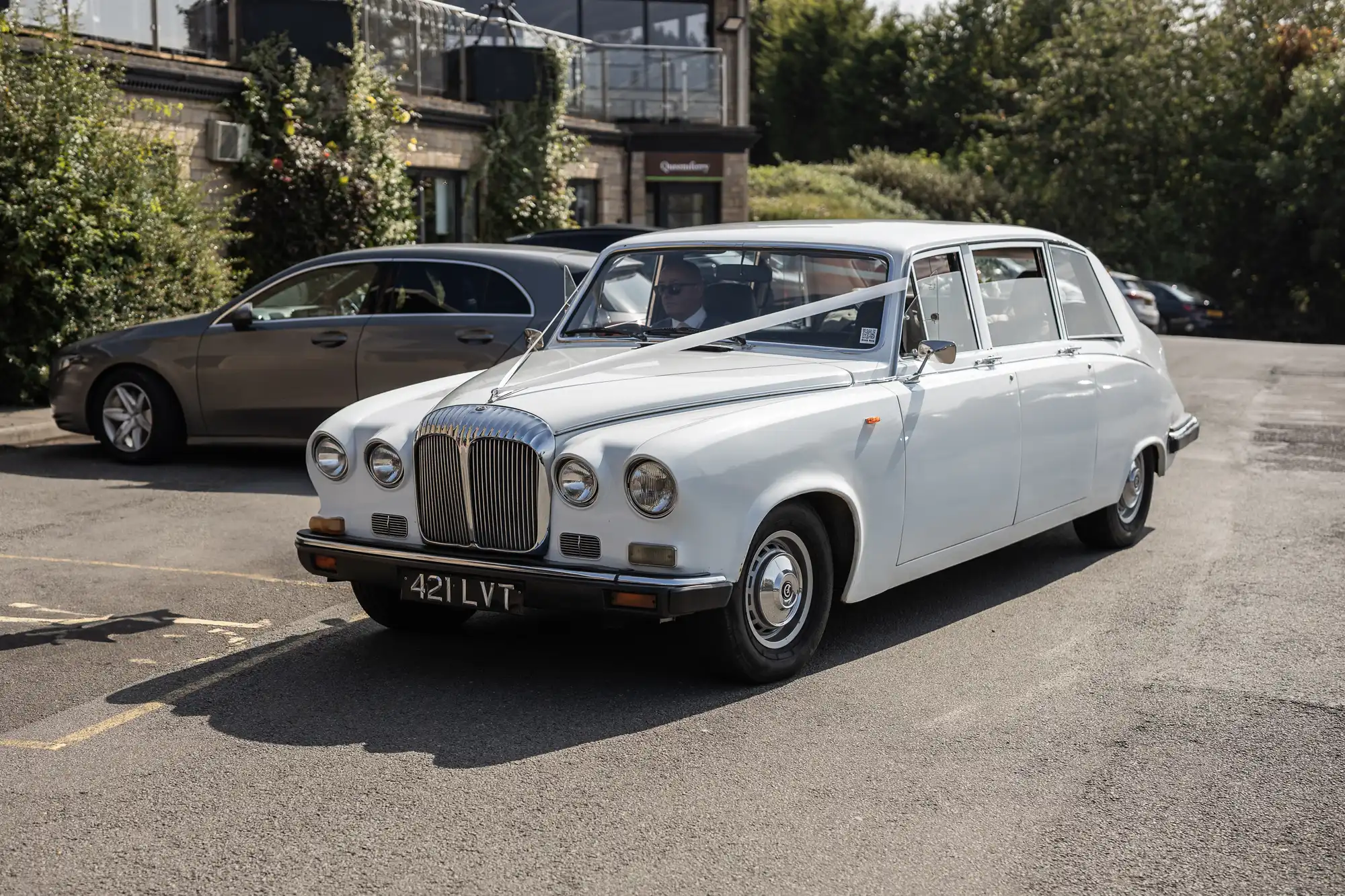 A classic white wedding car with a ribbon tied on the bonnet is parked on a sunlit street. The car has vintage styling and a number plate reading "421 LVT." Other vehicles and greenery are visible in the background.