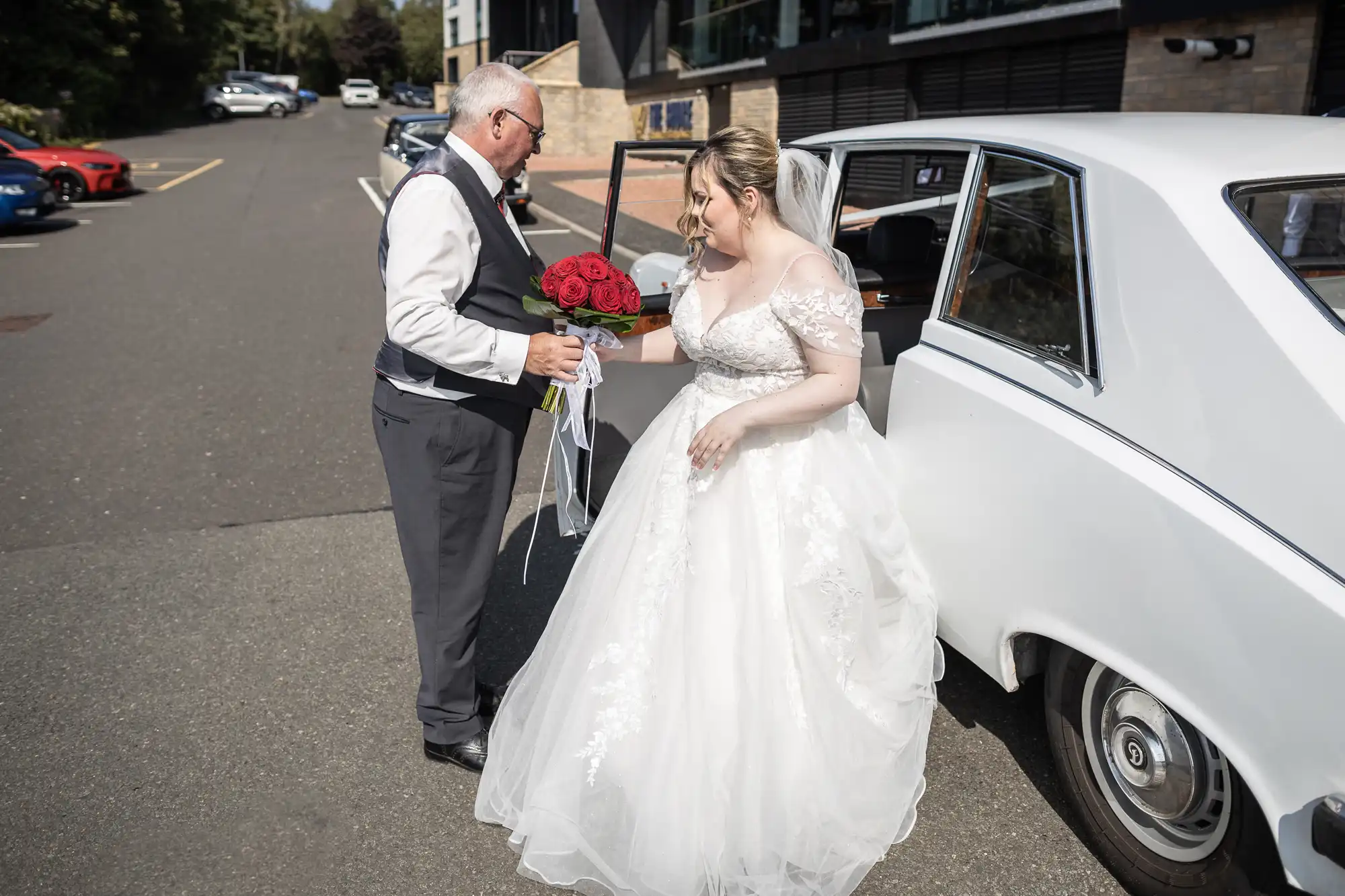 A bride in a white dress and veil steps out of a classic white car, assisted by an older man in a waistcoat and tie who holds a bouquet of red roses. They are in a car park near a building, and several colourful cars are parked in the background.