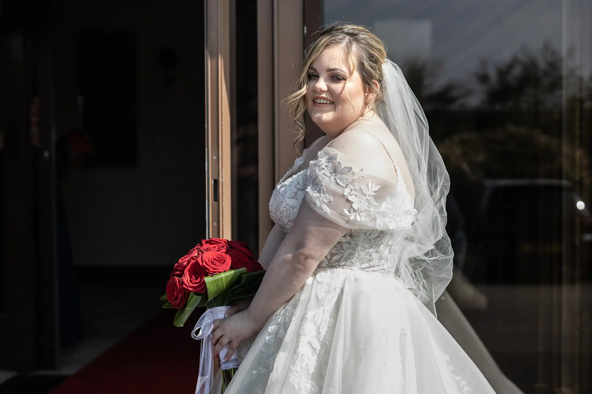A smiling bride in an off-the-shoulder white lace wedding gown and veil stands outside a building, holding a bouquet of red roses with white ribbon. Sunlight highlights her face and dress as she looks over her shoulder towards the camera.