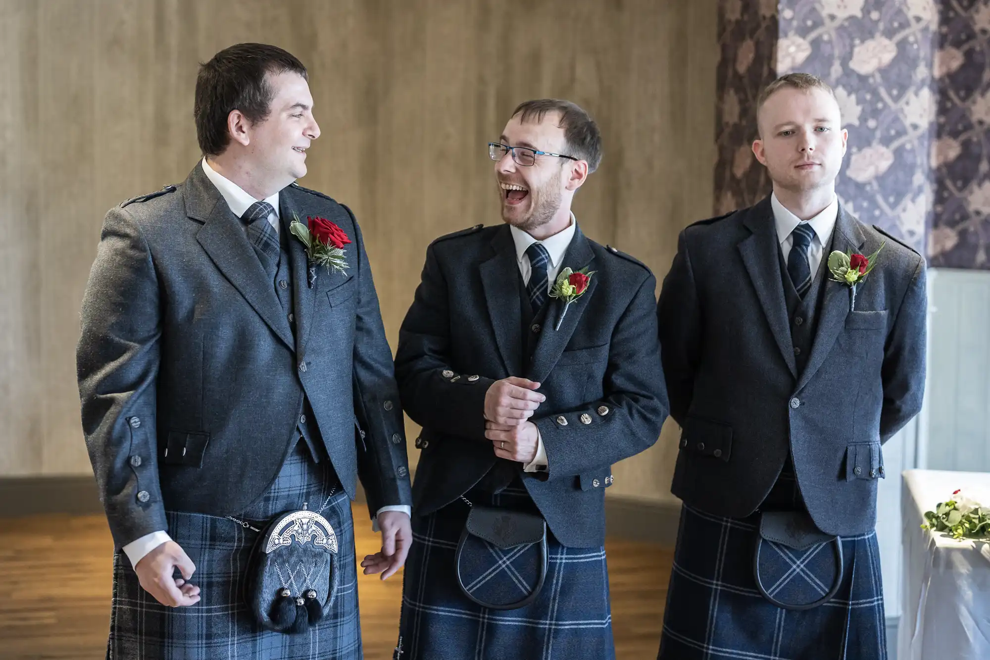 Three men in dark tartan kilts and matching jackets stand indoors. Two are smiling and laughing, while one stands more serious. Each wears a buttonhole with a red rose on their jacket lapel. The background features muted walls and soft lighting.